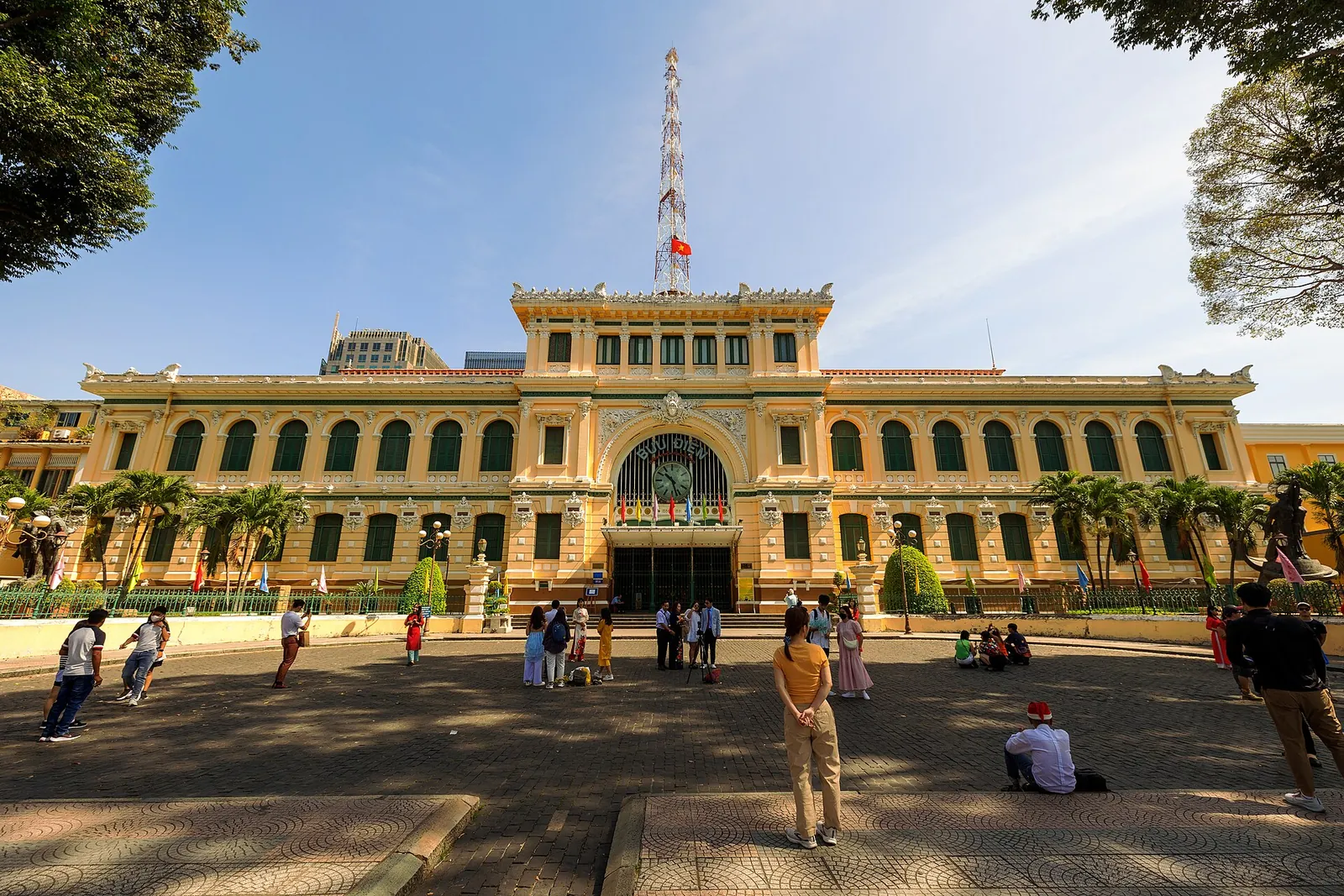 Yellow facade of the Saigon Central Post Office with its arched clock entrance under a blue sky, visitors on the forecourt