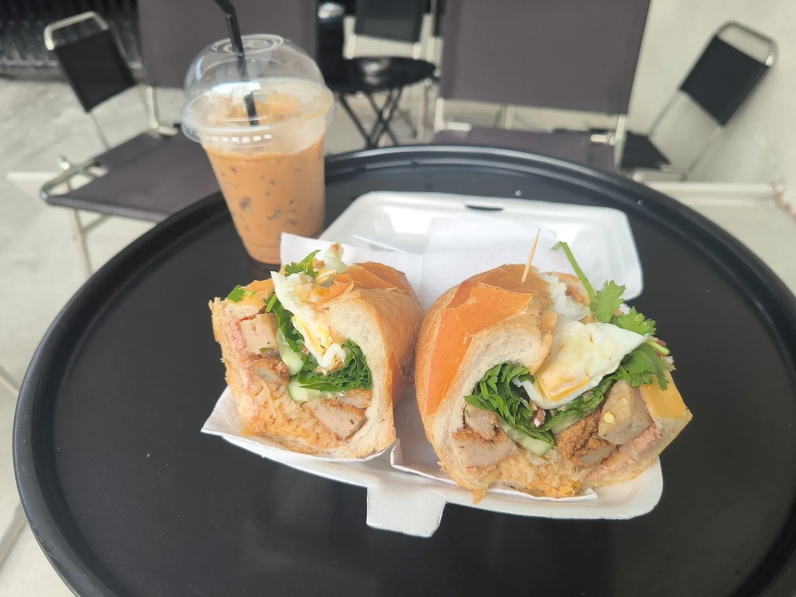 A banh mi cut in two halves, filled with meat, fried egg, herbs and pickles in a takeaway box, with an iced milk coffee behind