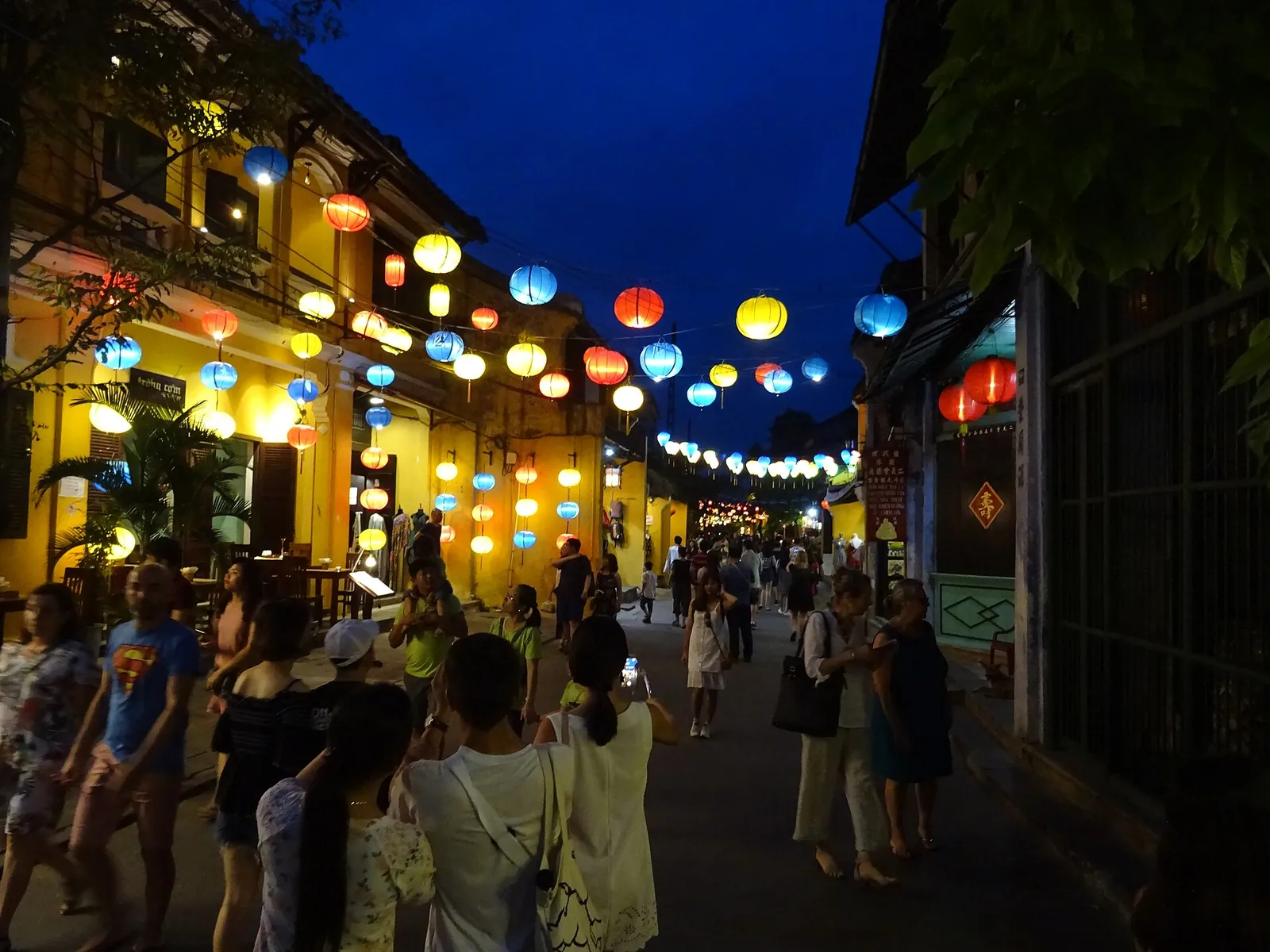 Crowds walking a lantern-lit street of Hoi An's Old Town at dusk, with coloured lanterns strung overhead