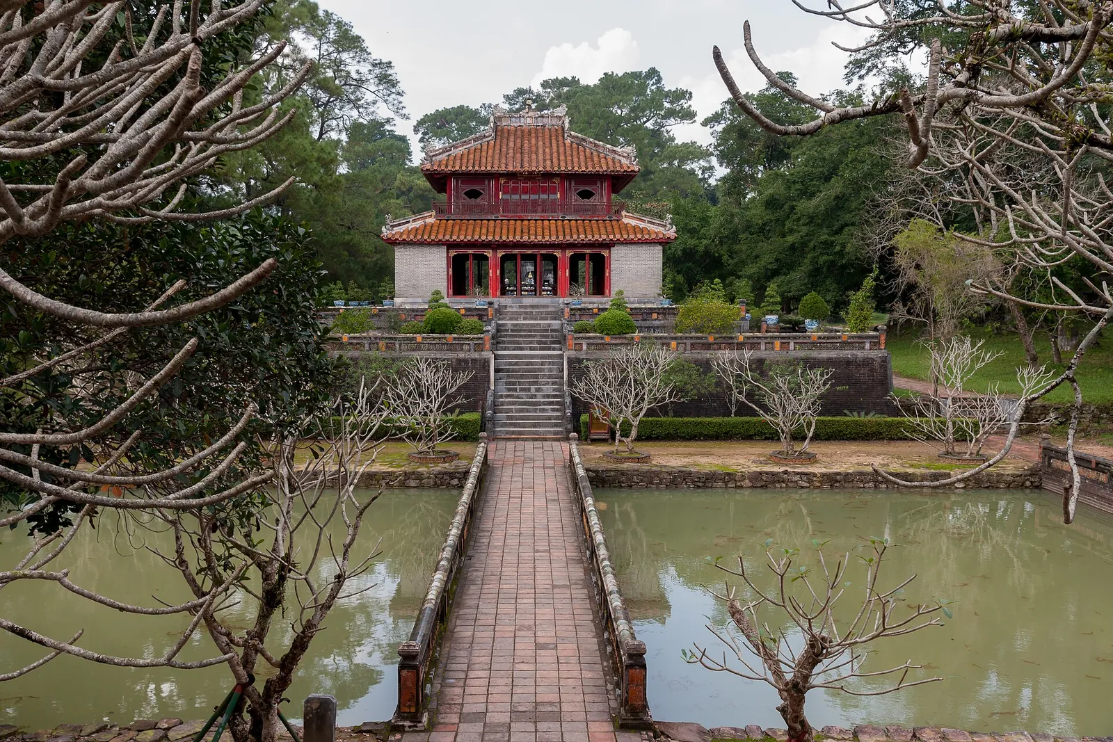 A stone causeway crossing a pond to a red and yellow pavilion at the tomb of Emperor Minh Mang