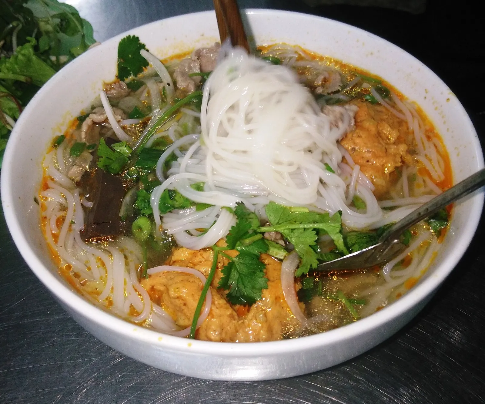 A bowl of bun bo Hue with round rice noodles, beef, pork and congealed blood in a red-orange broth with herbs
