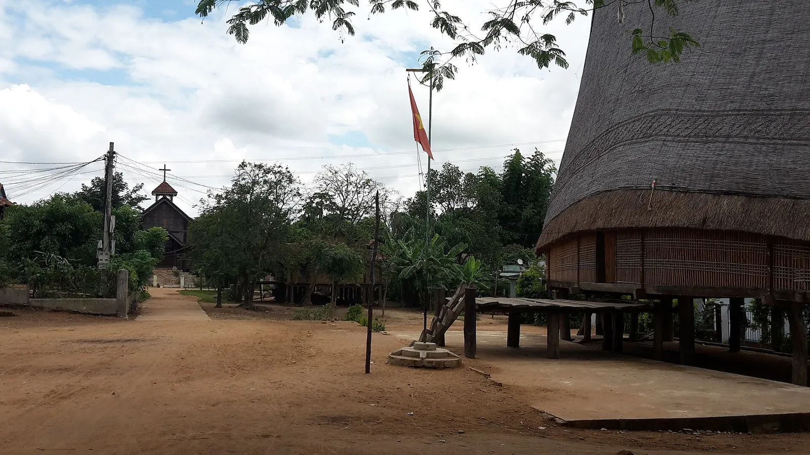 A wide dirt lane in the Ba Na village of Kon K'tu, with the thatched roof and stilts of a rong house on the right