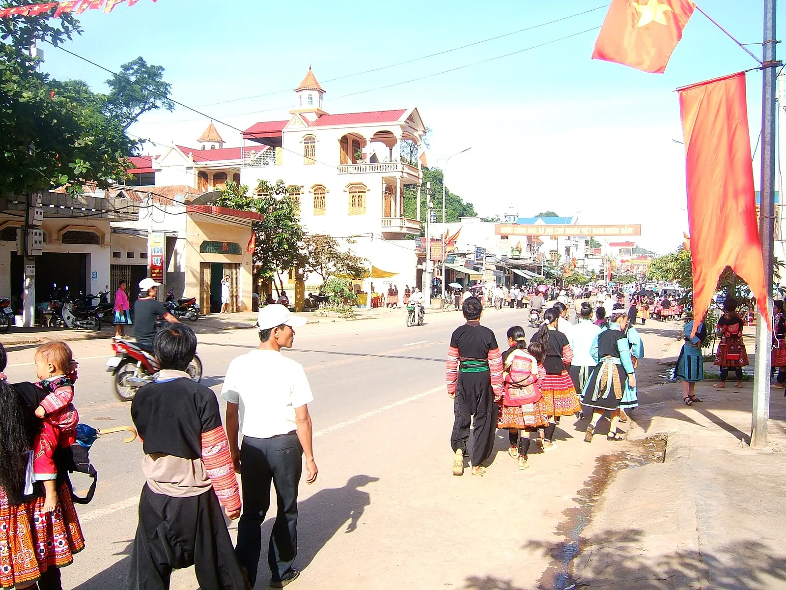 People in Hmong dress walking along the main street of Moc Chau town under red flags