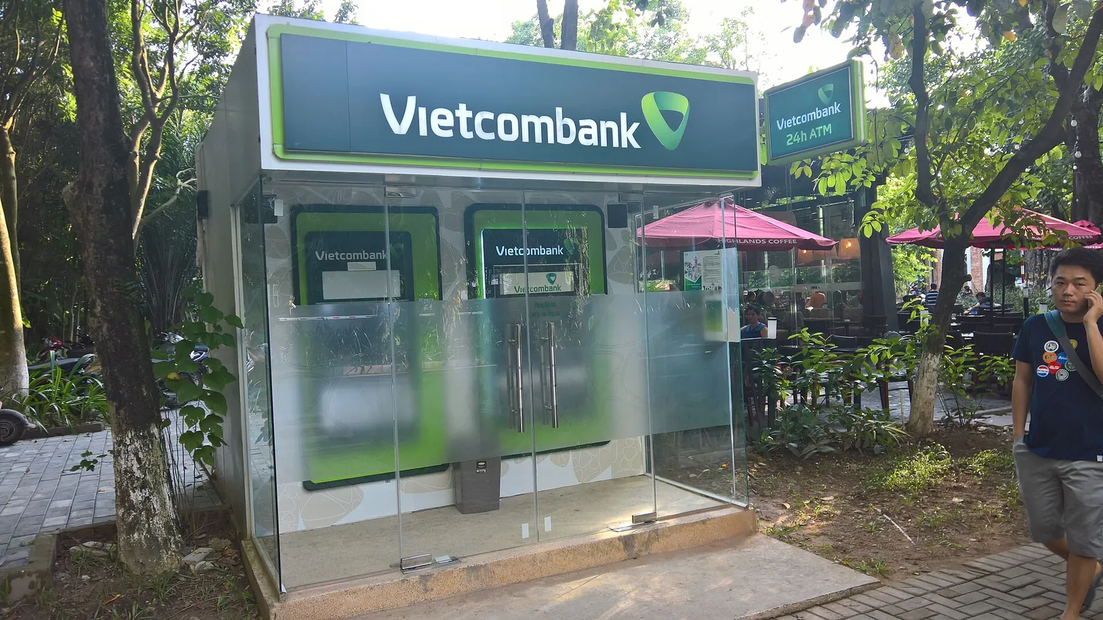 A glass Vietcombank ATM booth with two machines under trees beside a café