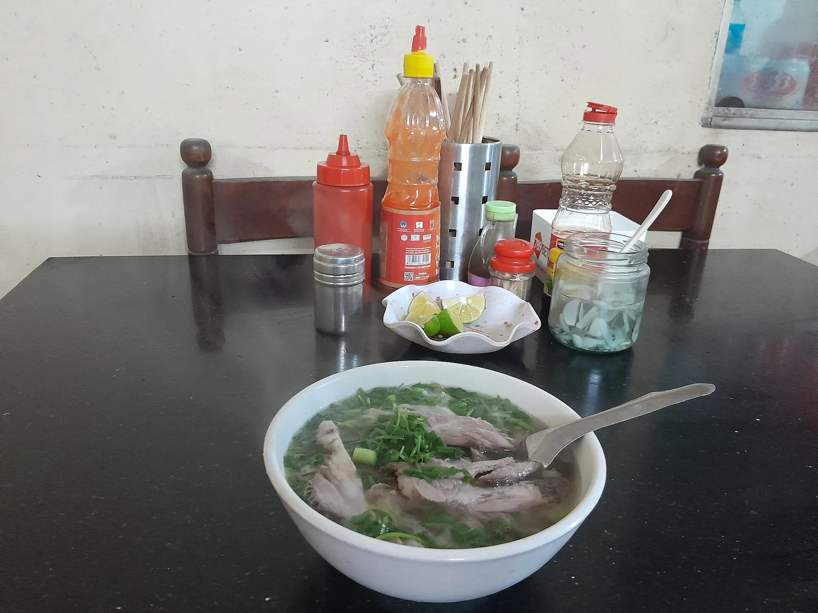 Bowl of beef phở with herbs on a table with chilli sauce, lime and condiments