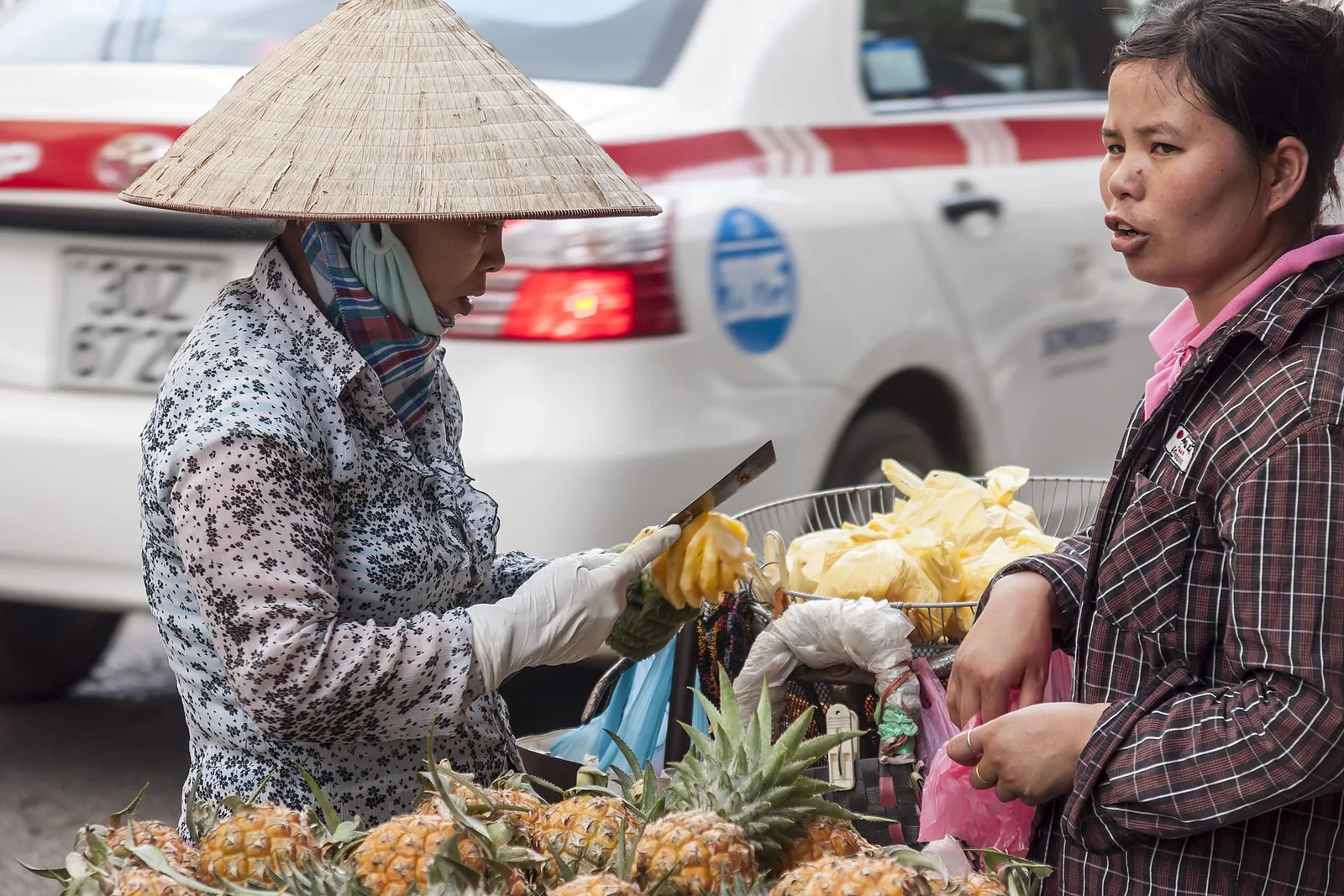 Vendor in a conical hat cutting pineapple for a customer beside a taxi in Hanoi