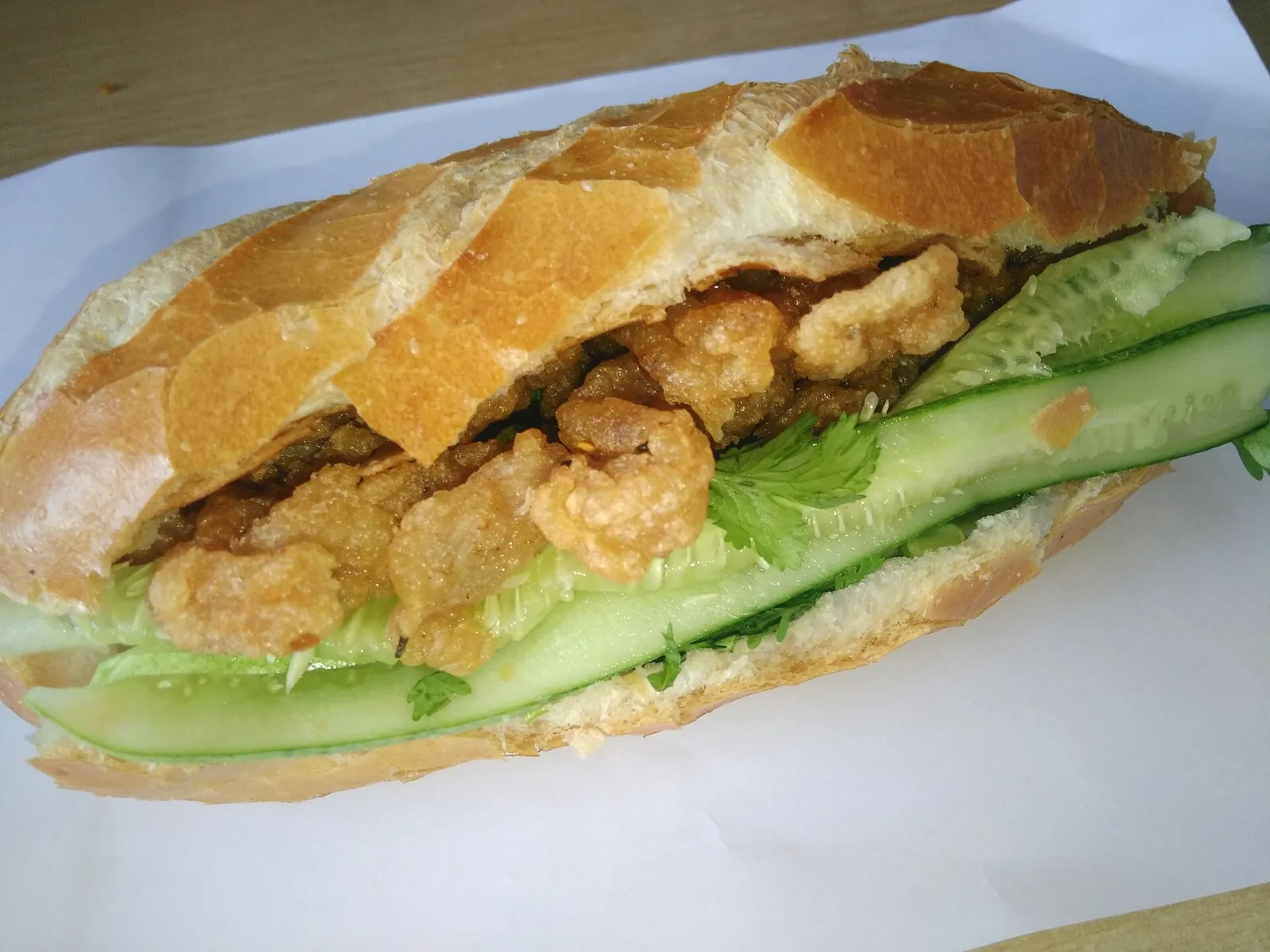 A baguette filled with fried fish cake, cucumber and herbs on white paper