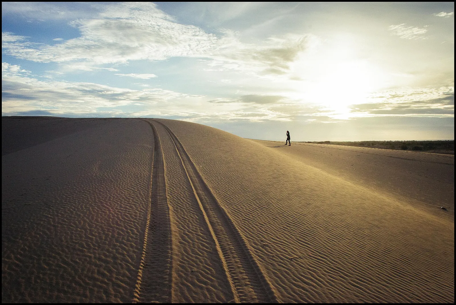 A lone walker on the crest of the white sand dunes near Mui Ne, with tyre tracks in the sand and the sun low in the sky