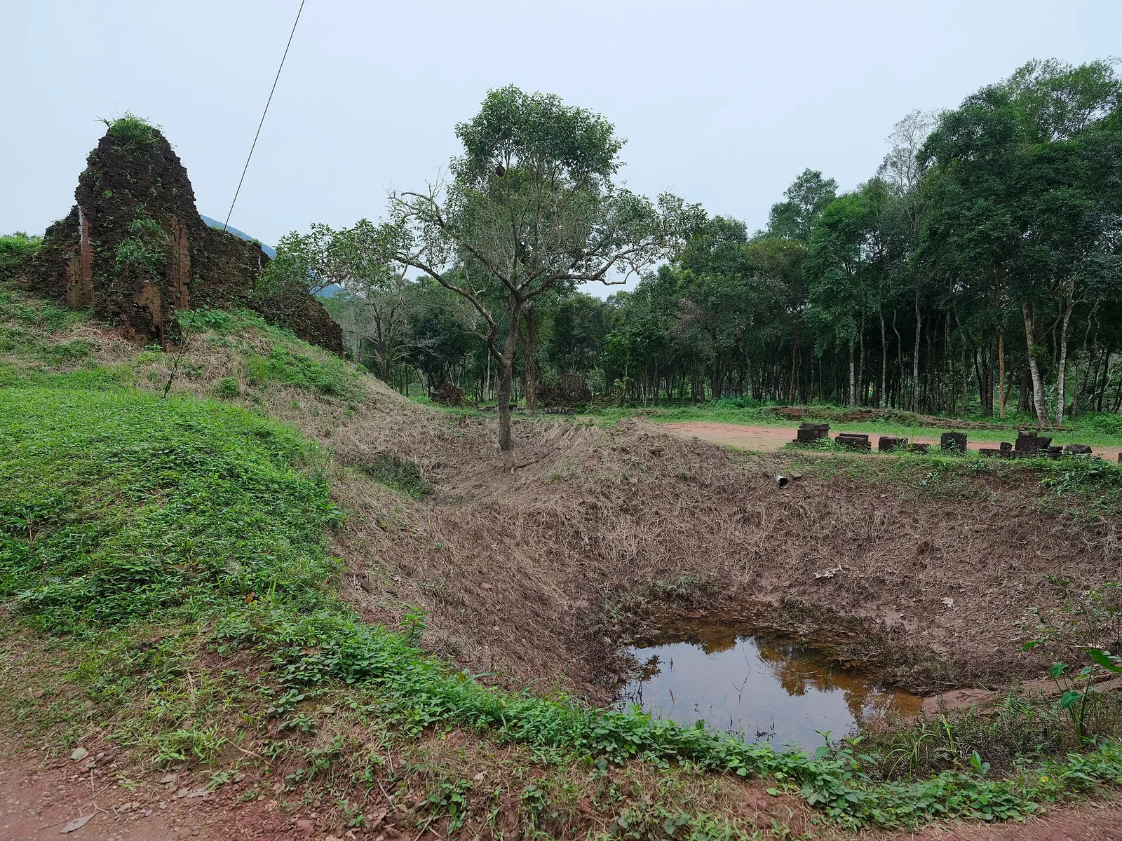 Grassy bomb crater with a pool of water beside ruined towers at My Son