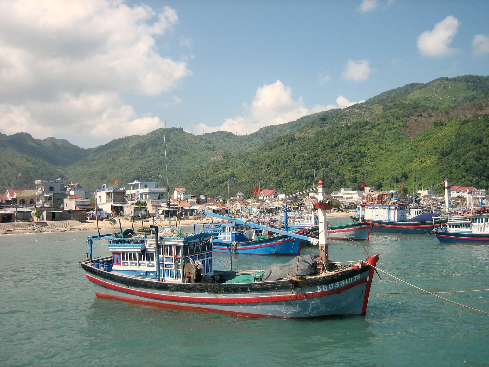 Blue and red wooden fishing boats moored in front of a fishing village and green hills on Nha Trang Bay