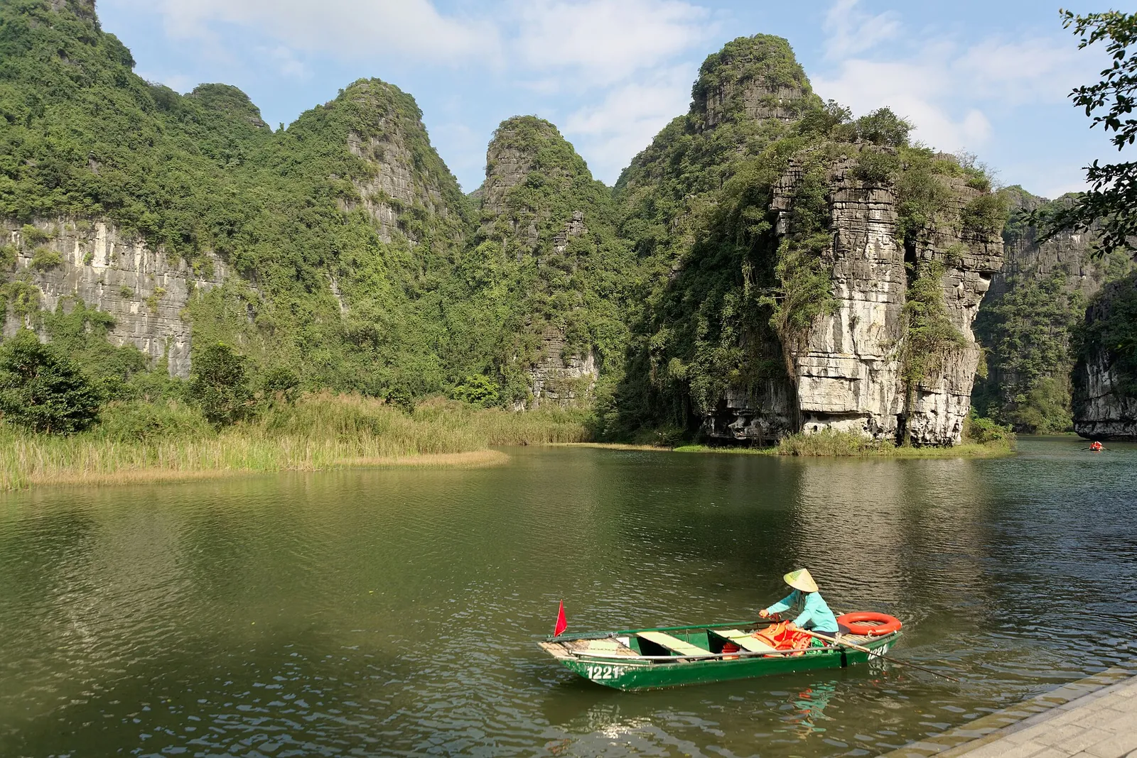 A rower in a small boat on a lake below green limestone cliffs at Trang An