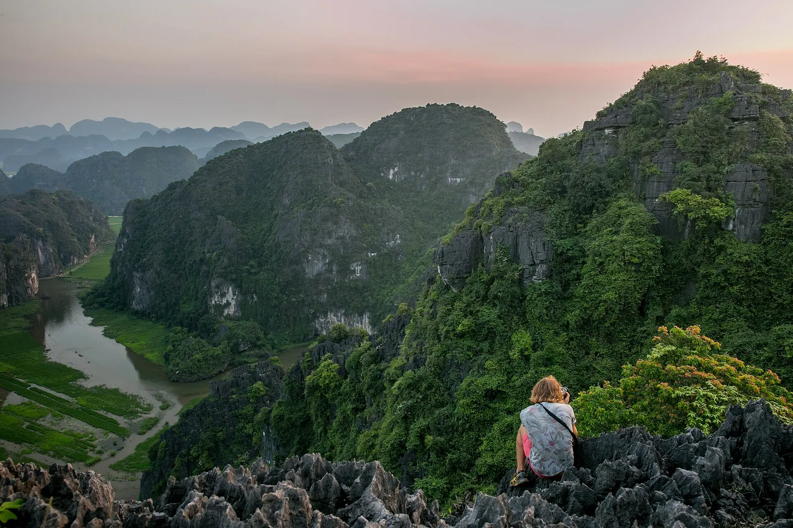 A visitor sitting on rocks at the top of Hang Mua above a river and rows of limestone peaks