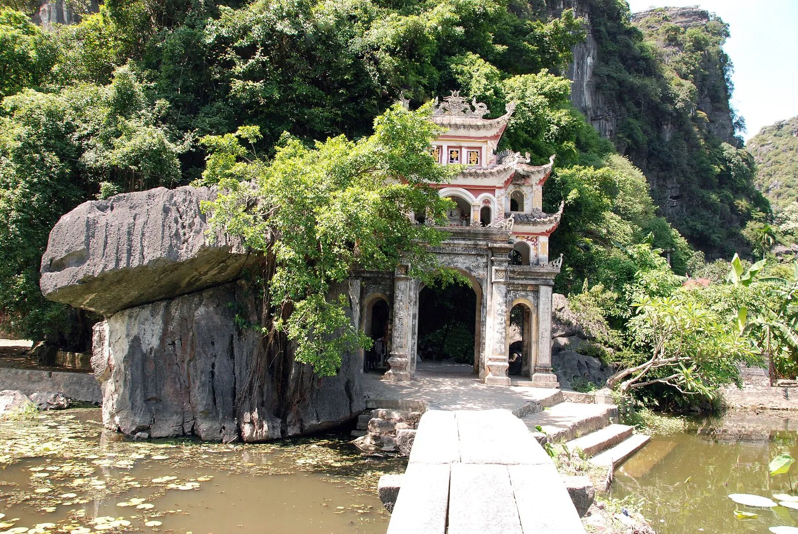 A stone bridge over a lotus pond leading to the gate of Bich Dong Pagoda below a cliff