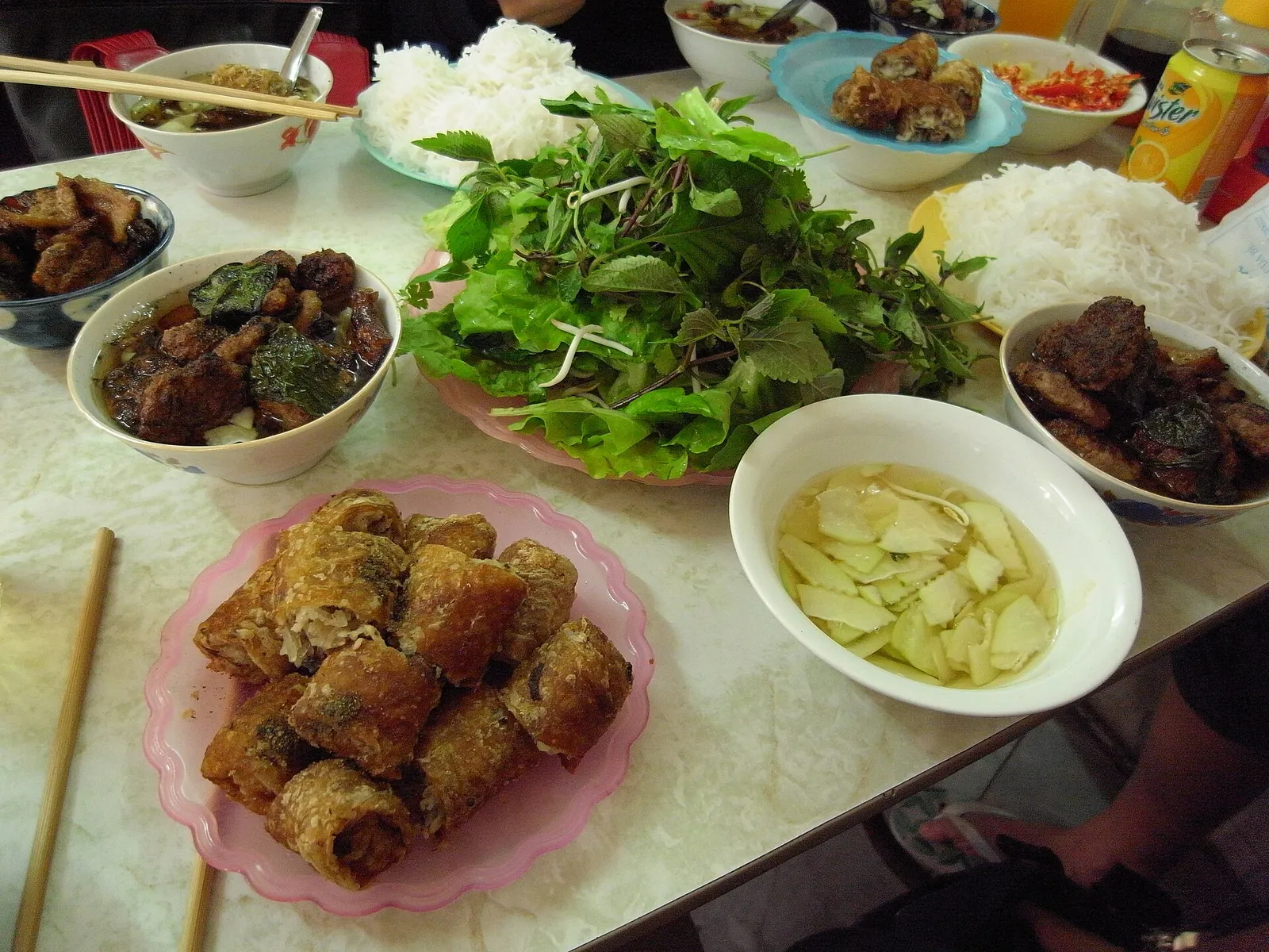A table of bun cha with grilled pork in dipping broth, rice noodles, fresh herbs and fried spring rolls