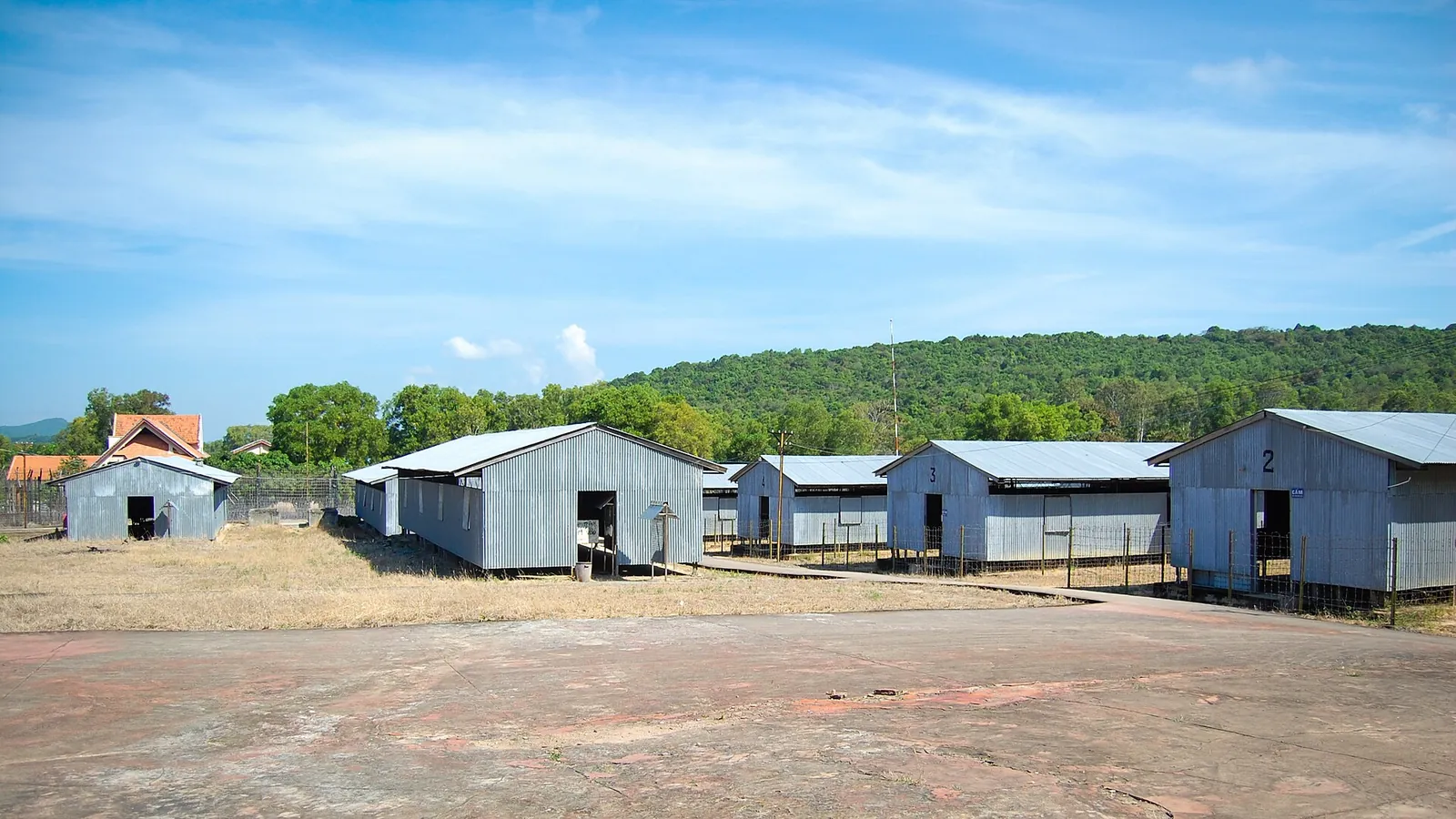 Rows of corrugated-metal prison huts on open ground at the Phu Quoc Prison relic site, with forested hills behind