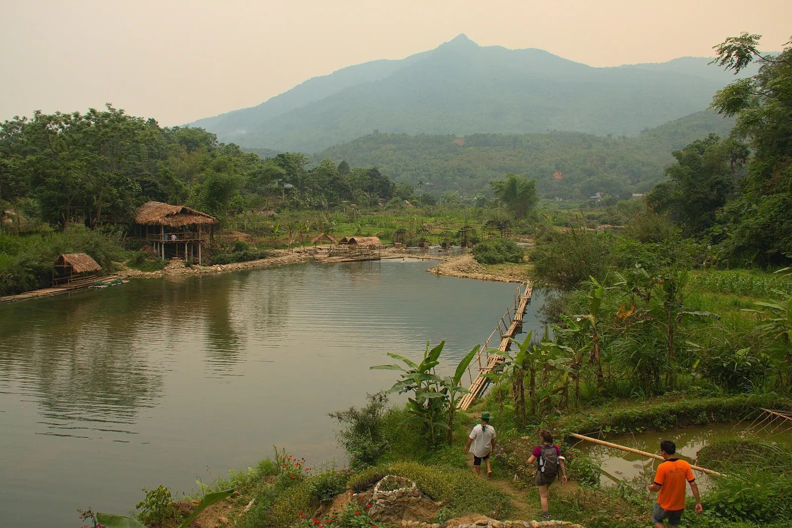 A pond fringed with trees and a wooden stilt house below a forested peak near Pu Luong