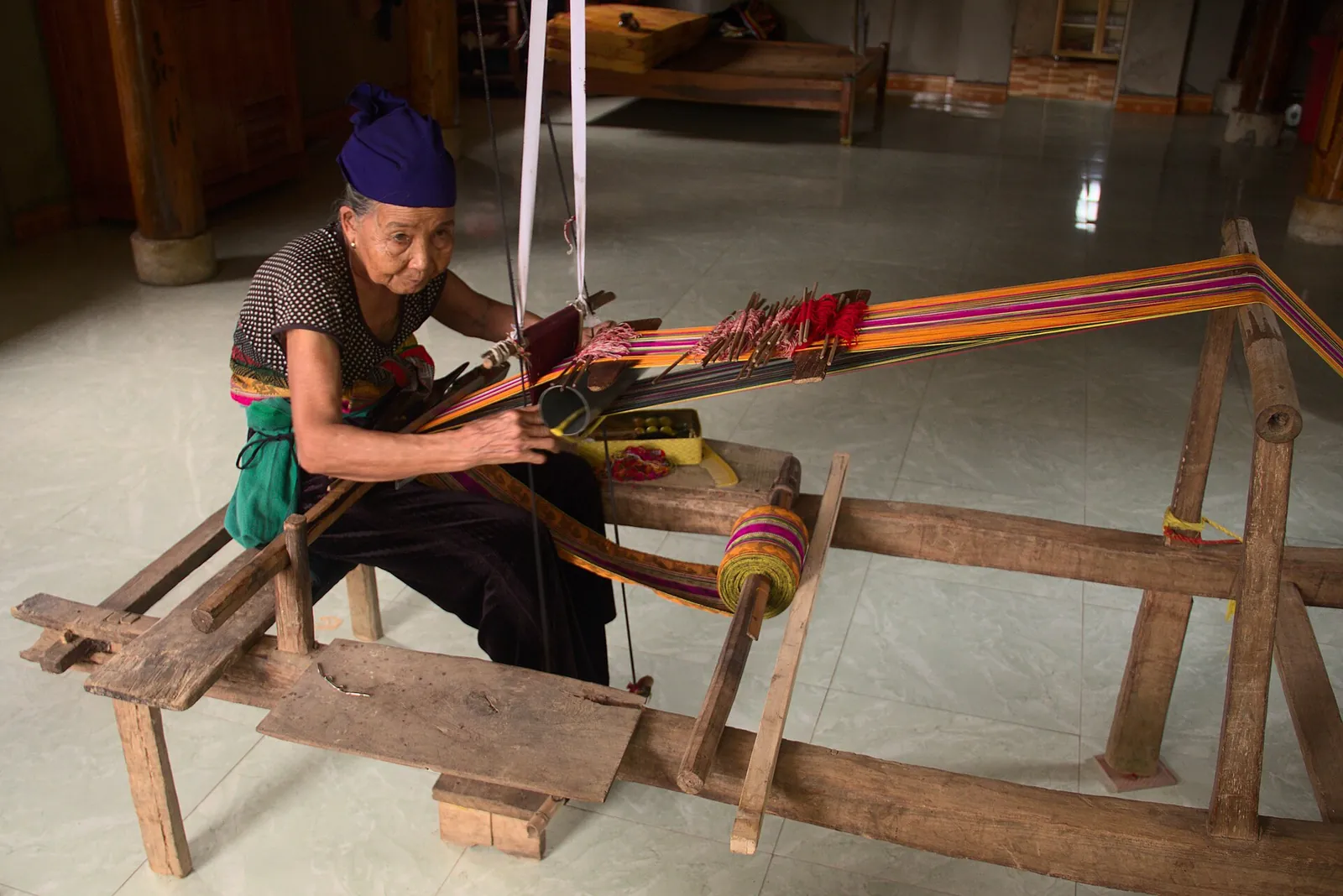 An older Thai woman in a headscarf weaving a striped textile on a wooden loom in Pu Luong