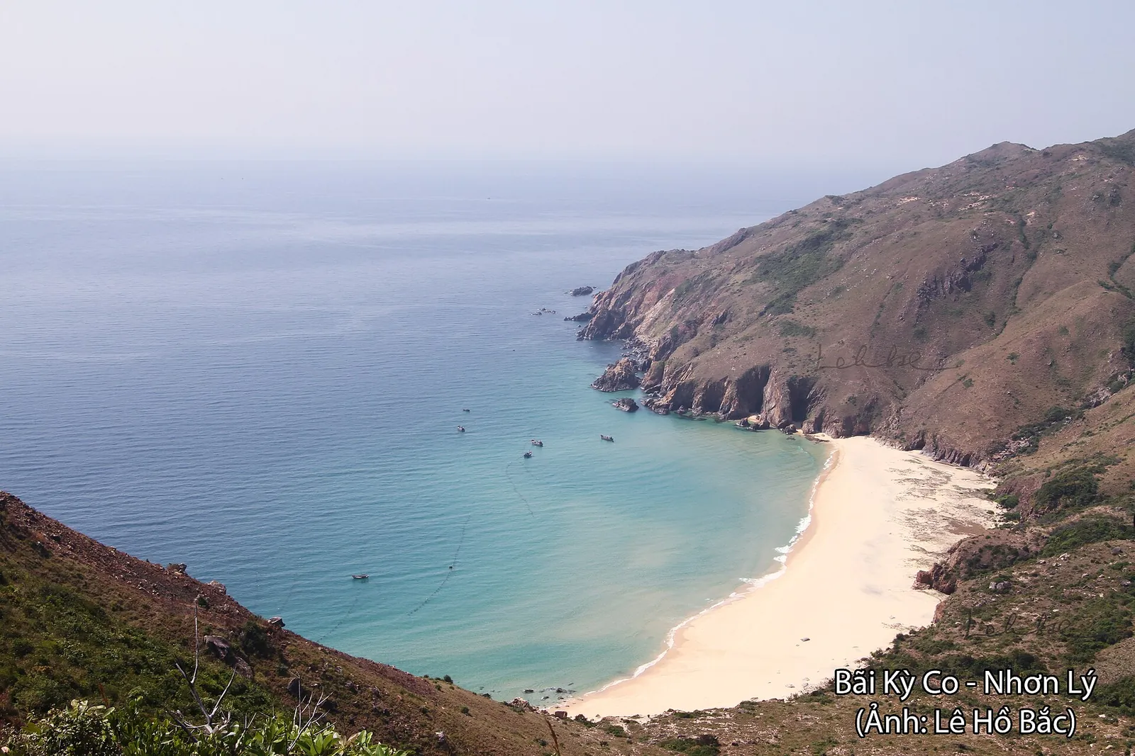 Ky Co beach, a white-sand cove below steep hills, with turquoise water and small boats in the bay
