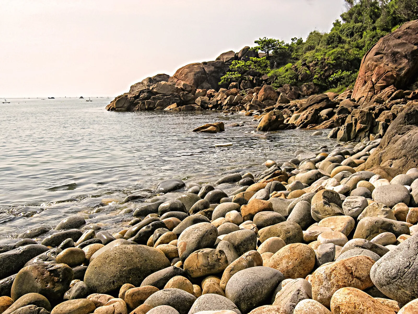 Rounded boulders and a beach of smooth egg-shaped stones beside a rocky, wooded headland