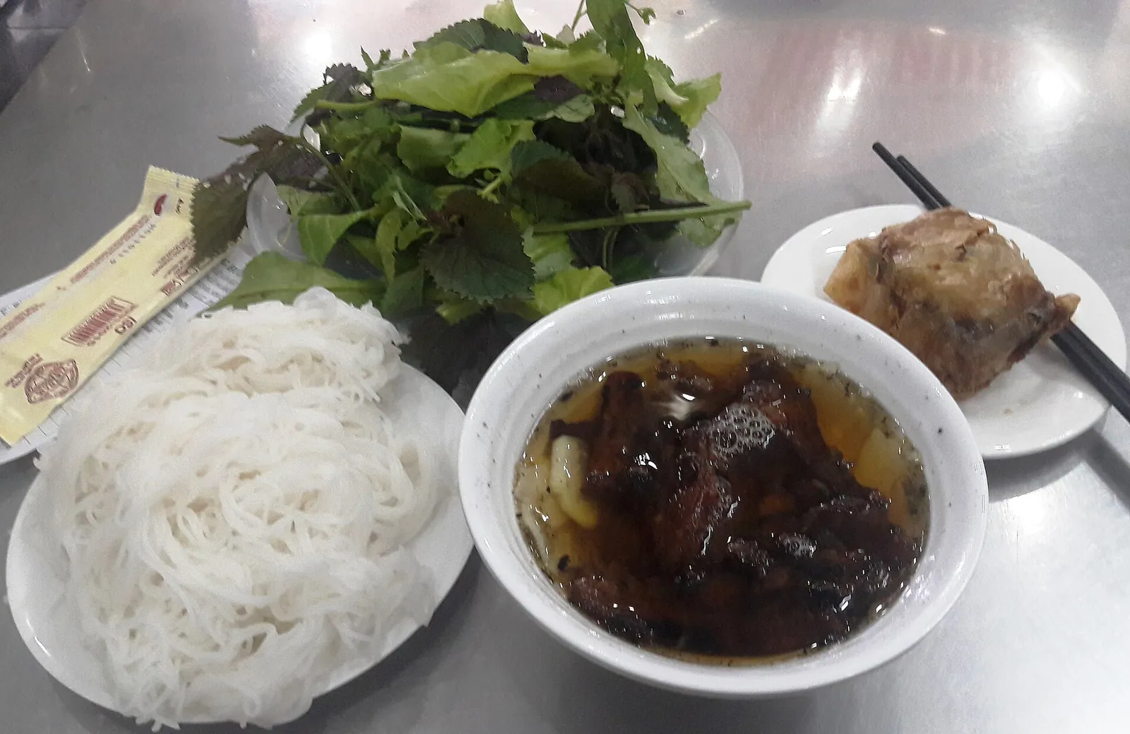 Bowl of bun cha with grilled pork in broth beside rice vermicelli, herbs and a spring roll