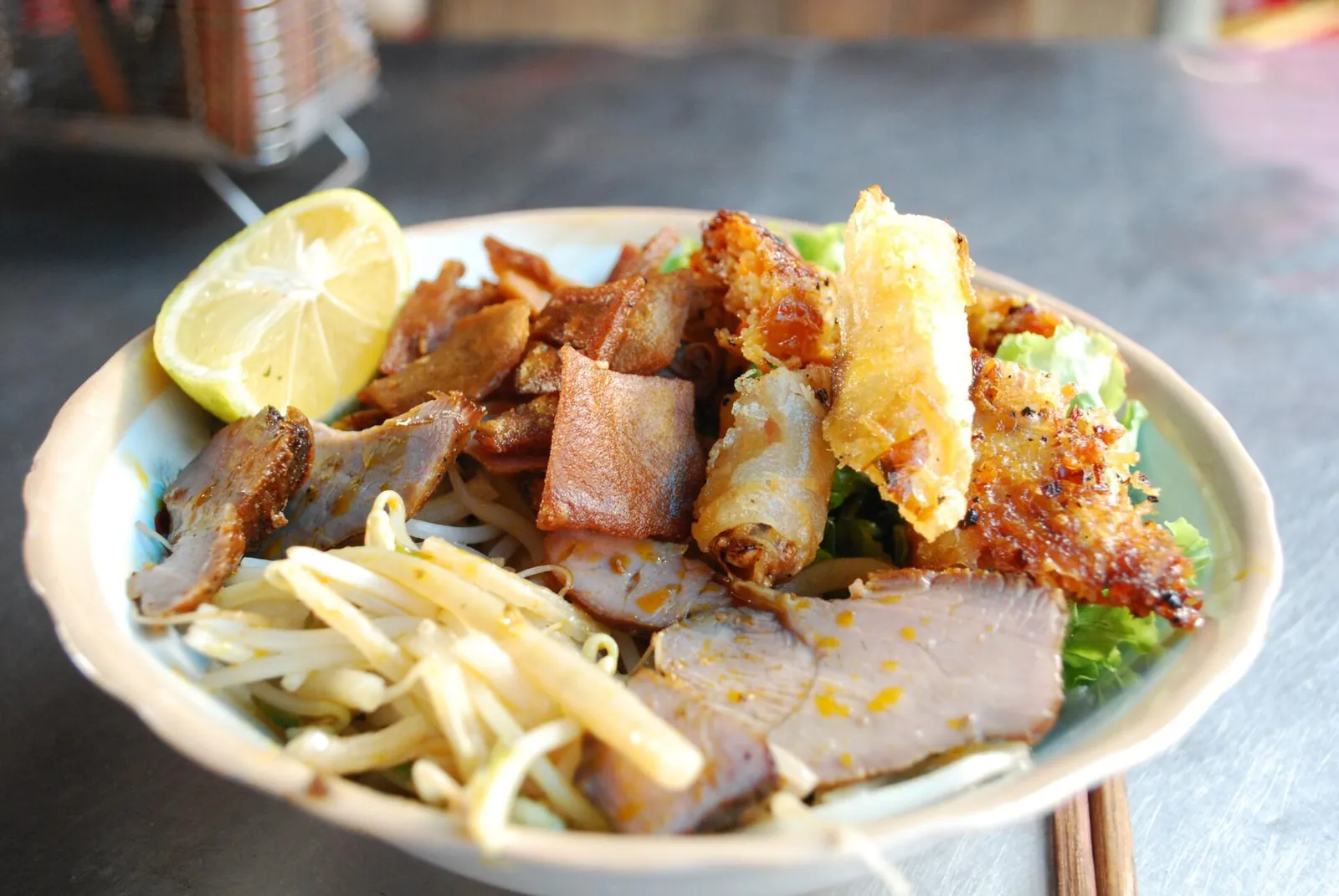 Plate of cao lau noodles with sliced pork, crisp crackling, fried spring rolls, bean sprouts and lime at a market stall in Hoi An