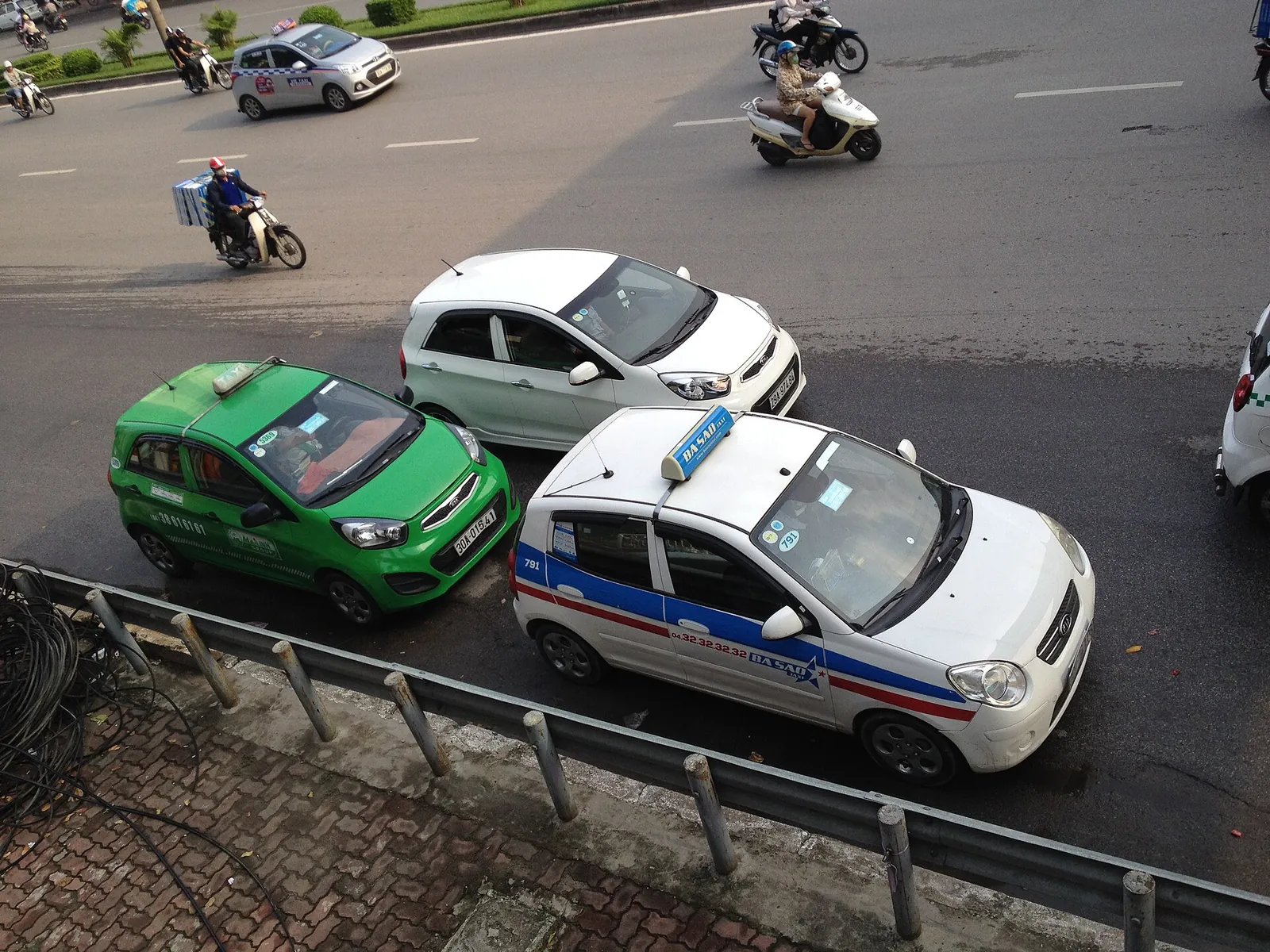 A green Mai Linh taxi and a white taxi on Đại Cồ Việt street in Hanoi, with motorbikes behind
