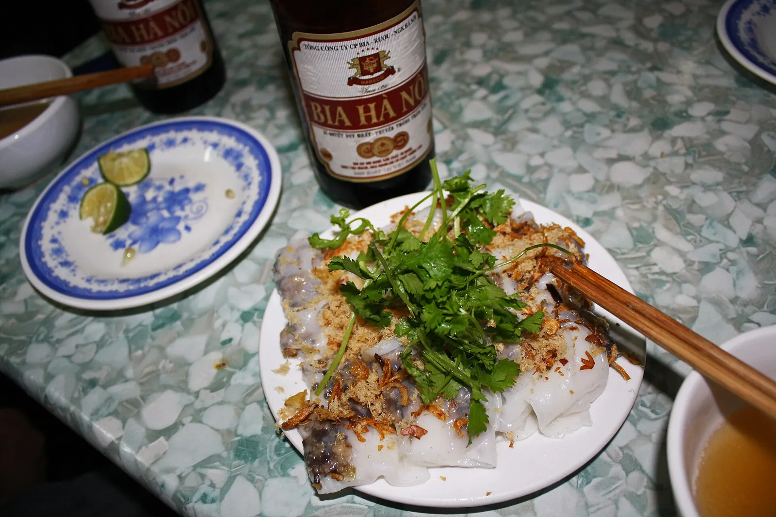 A plate of bánh cuốn steamed rice rolls topped with fresh herbs and fried shallots, with a bottle of Bia Hà Nội beside it