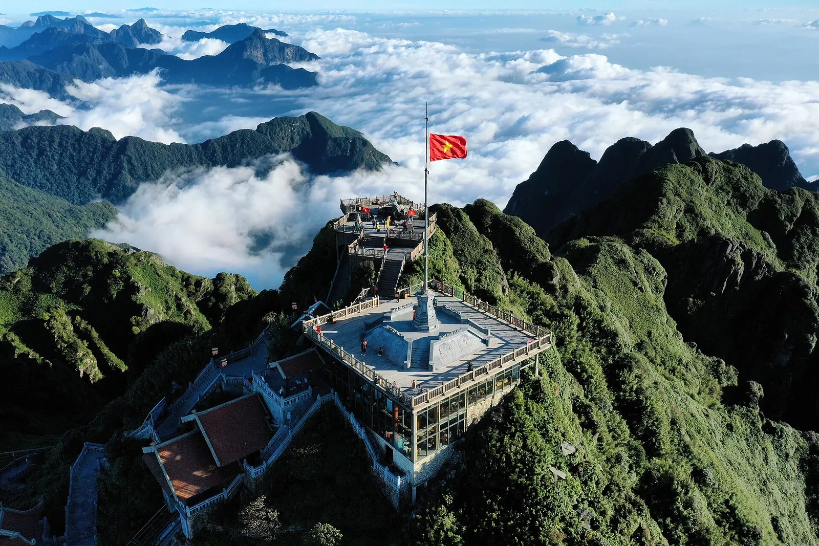 Aerial view of the Fansipan summit with a flag, viewing platform and pagoda roofs above a sea of clouds