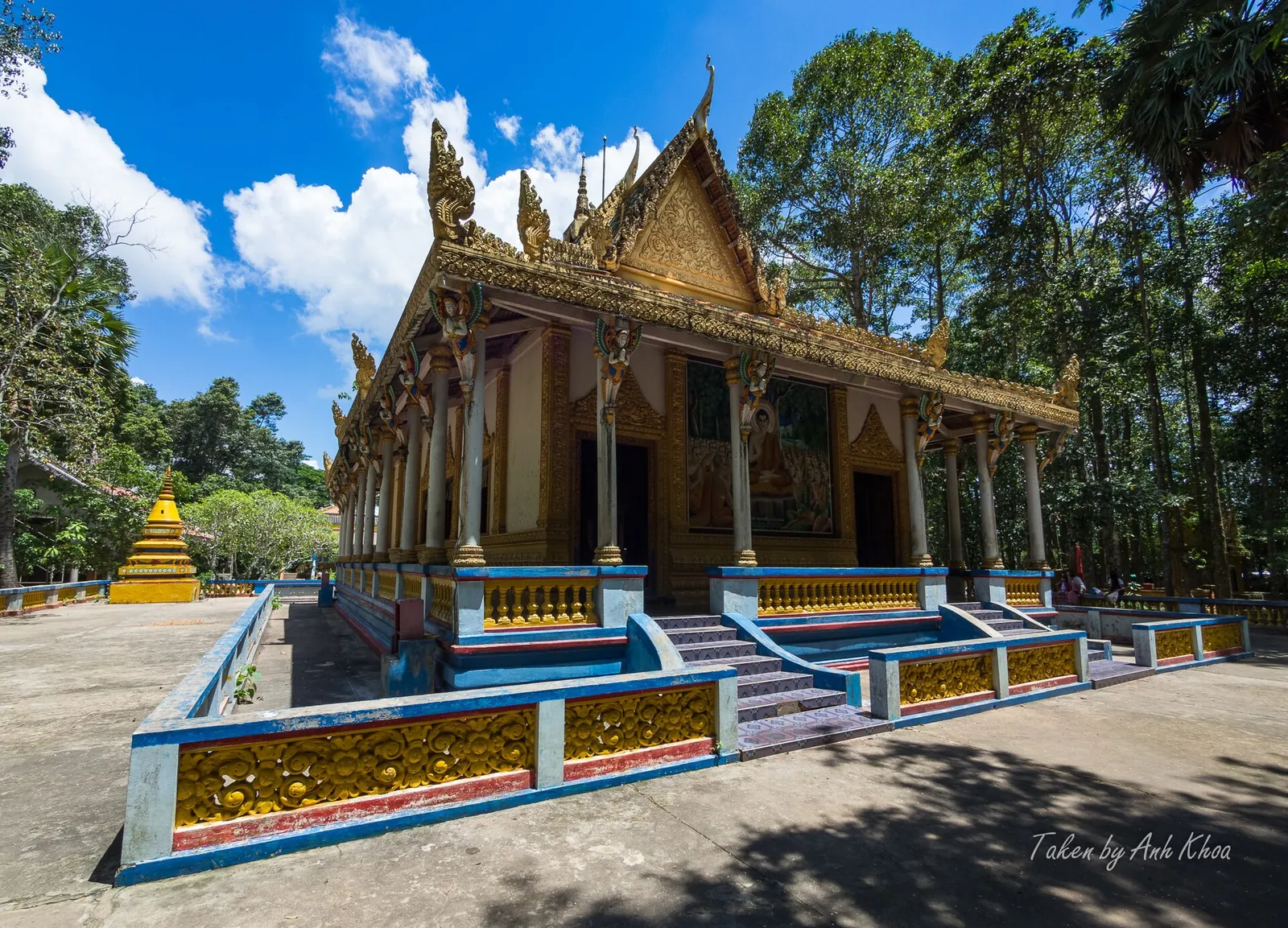 Gilded Khmer main hall of the Bat Pagoda in Soc Trang under a blue sky, surrounded by tall trees