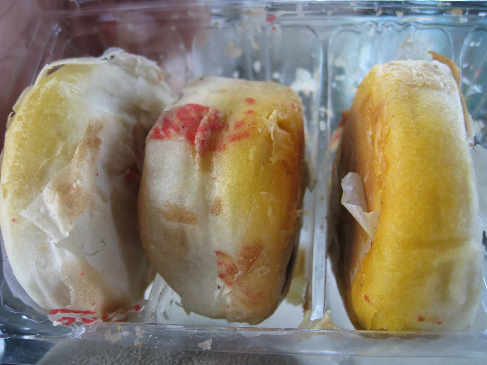Three bánh pía pastries with flaky white crusts and red stamps in a plastic tray