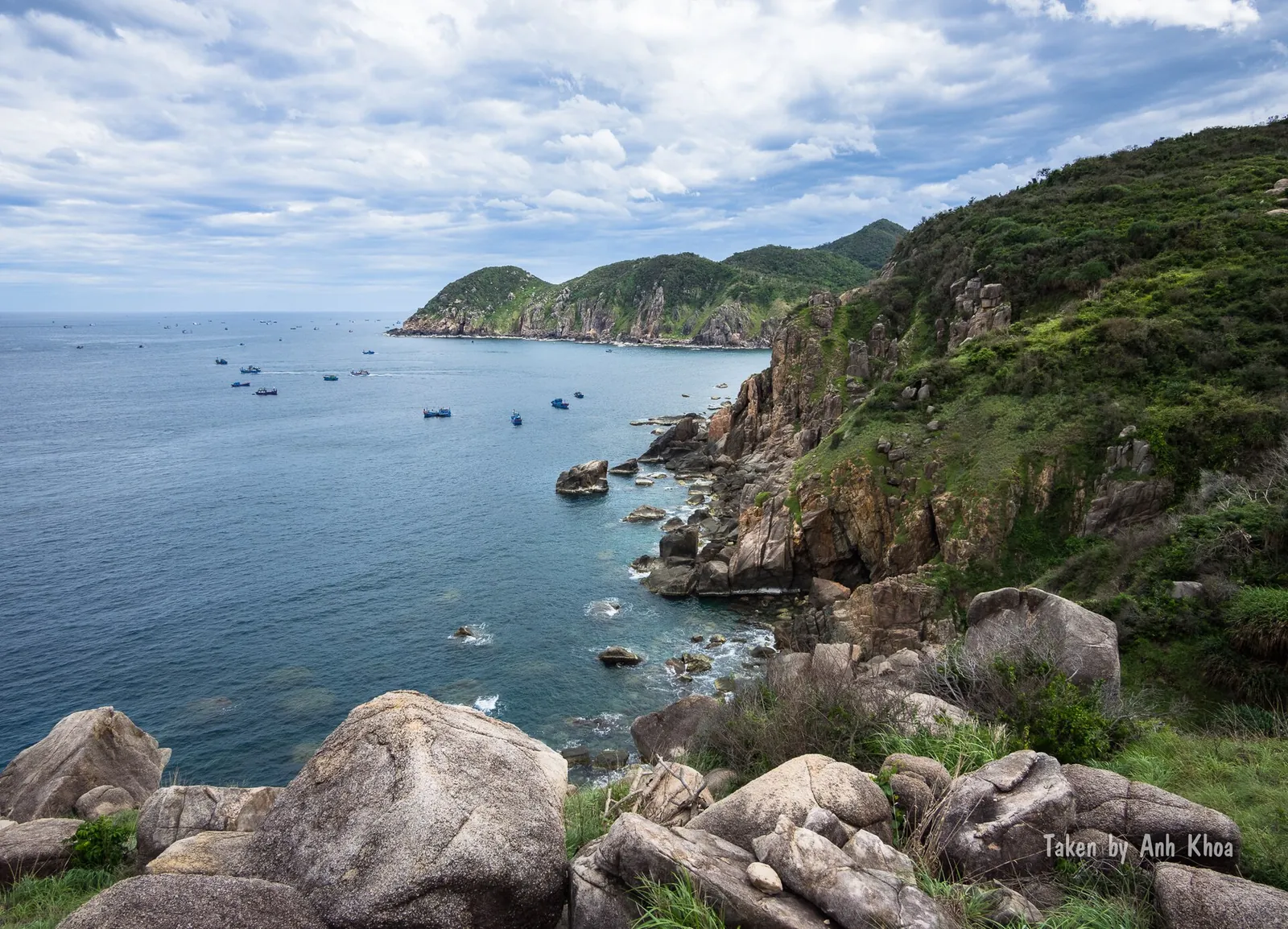 Boulders and a green rocky headland above a blue bay dotted with fishing boats at Mui Dien