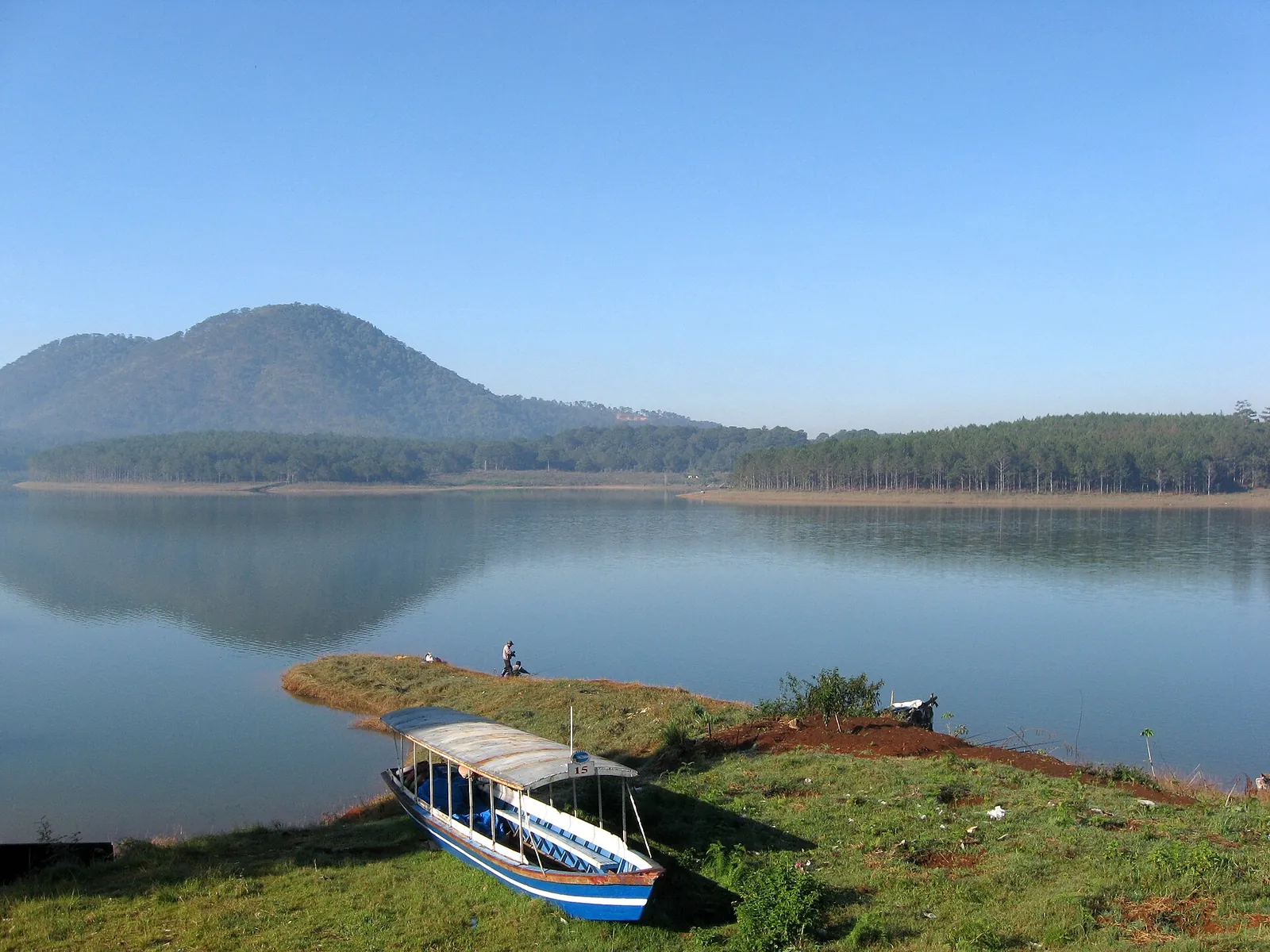 A covered wooden boat moored on the grassy shore of Tuyen Lam Lake with a forested hill behind