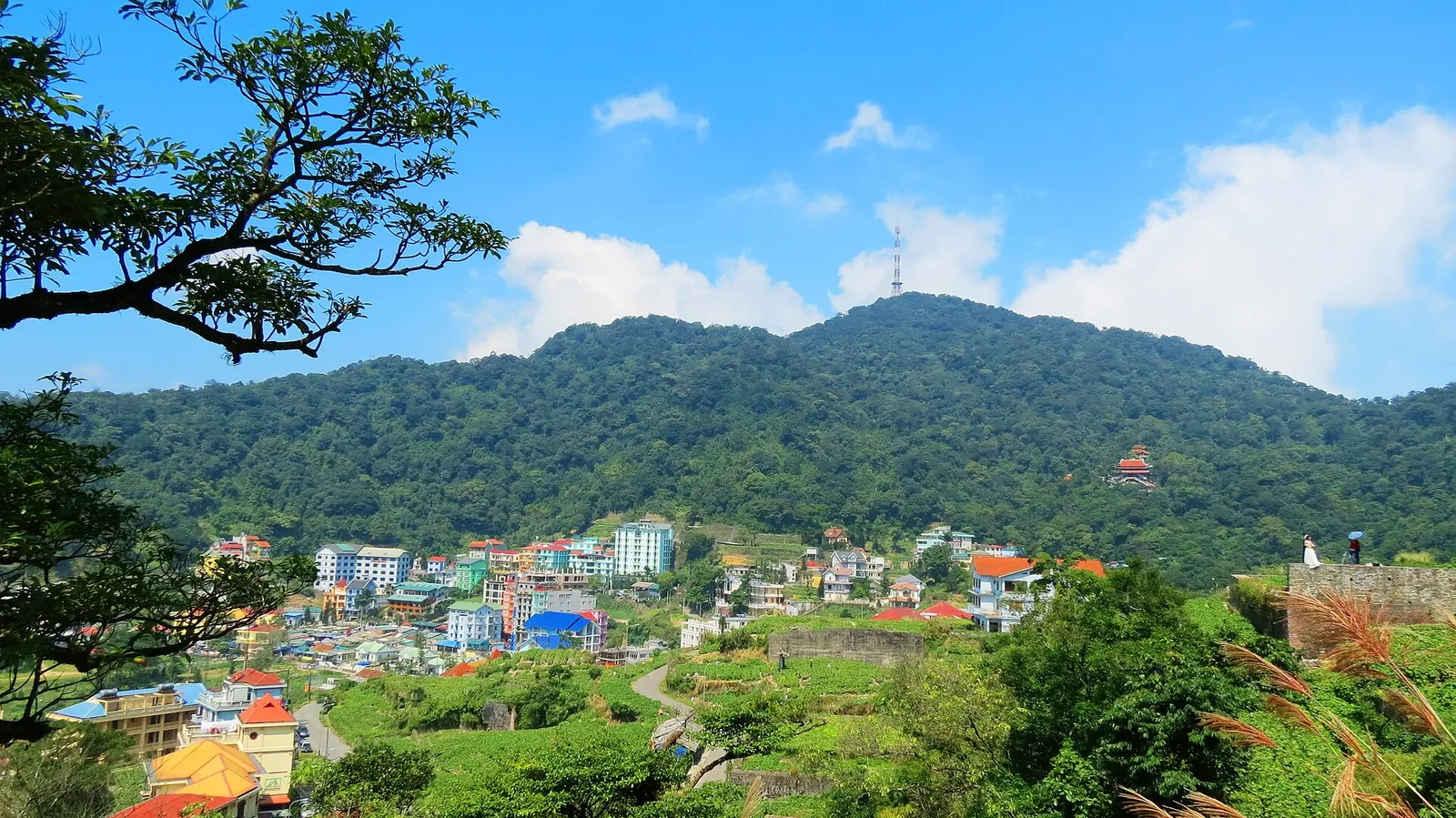 Hotels and villas of Tam Dao town on a green hillside below a forested ridge with a transmission mast, under a blue sky