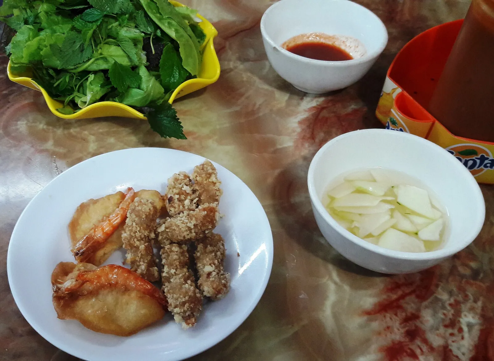 A plate of banh tom fritters with a dish of herbs, dipping sauce and sliced papaya