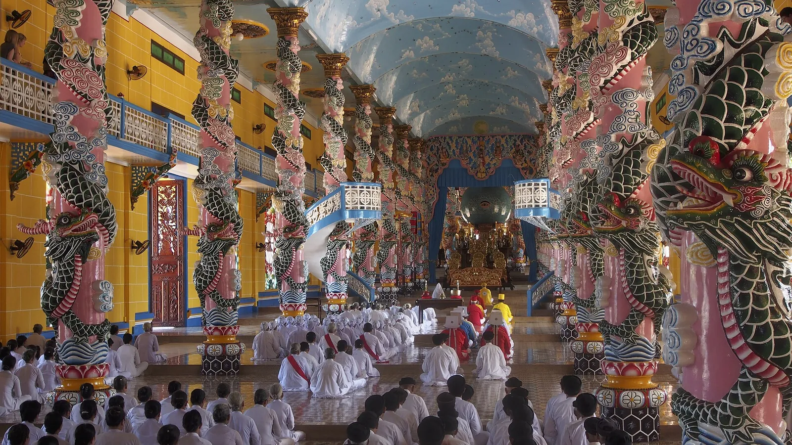 Rows of white-robed Cao Dai worshippers kneeling between dragon-wrapped columns inside the Tay Ninh Holy See