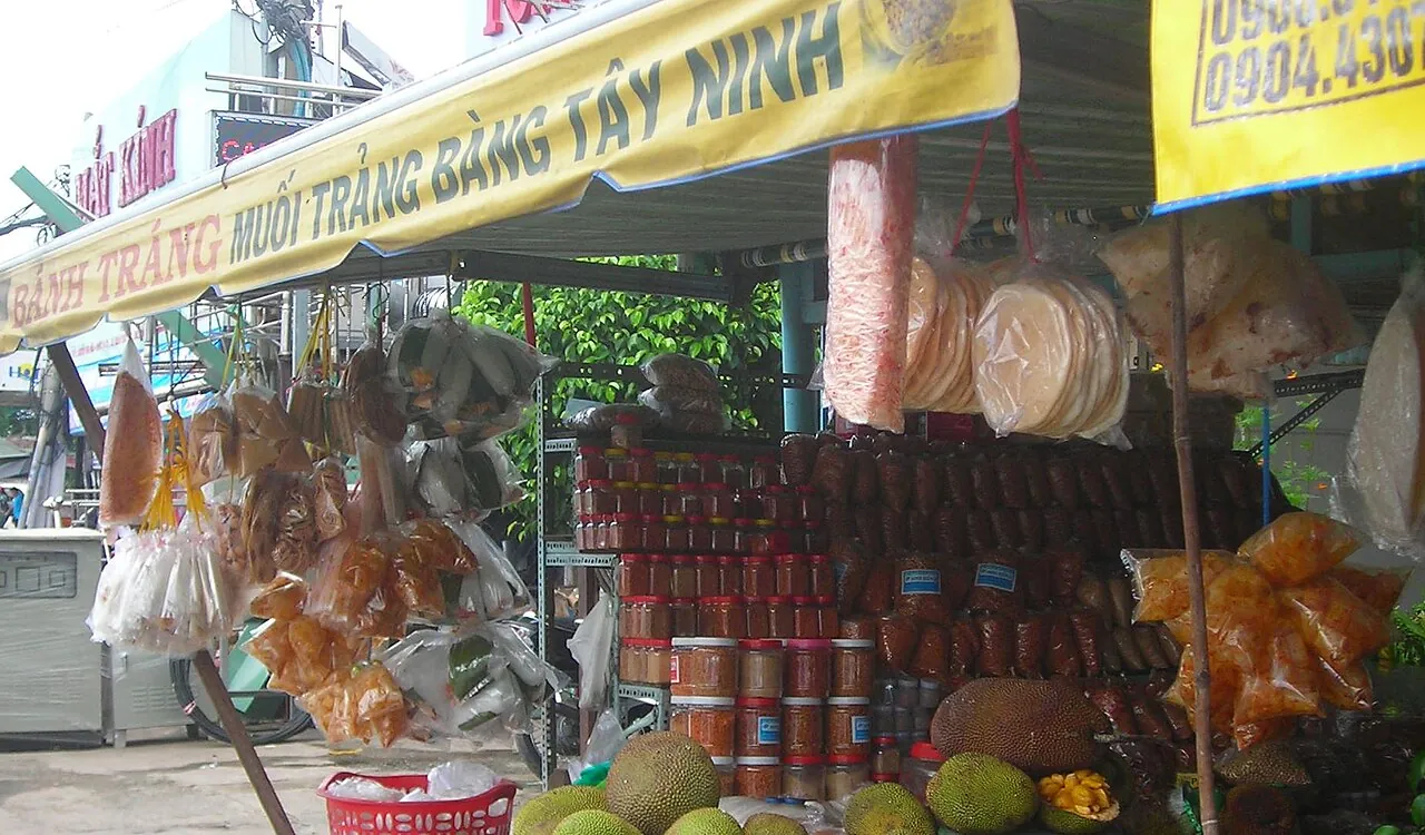 Roadside stall hung with bags of rice paper, jars of shrimp salt and fruit under a Trang Bang Tay Ninh sign