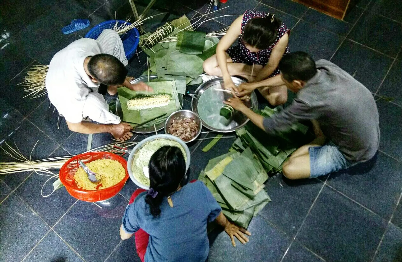 A family sitting on the floor wrapping banh tet sticky rice cakes in banana leaves before Tet