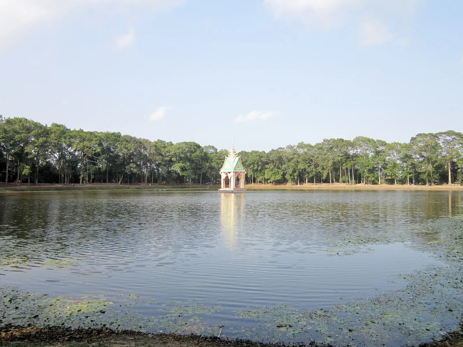 Ao Ba Om pond in Tra Vinh under a blue sky, with a small pavilion on the water and a line of tall trees on the far bank