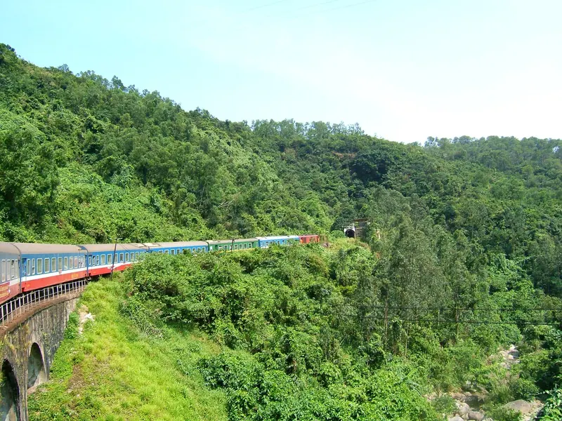 A blue and red Vietnam Railways passenger train crossing a bridge through green forested hills near the Hai Van Pass