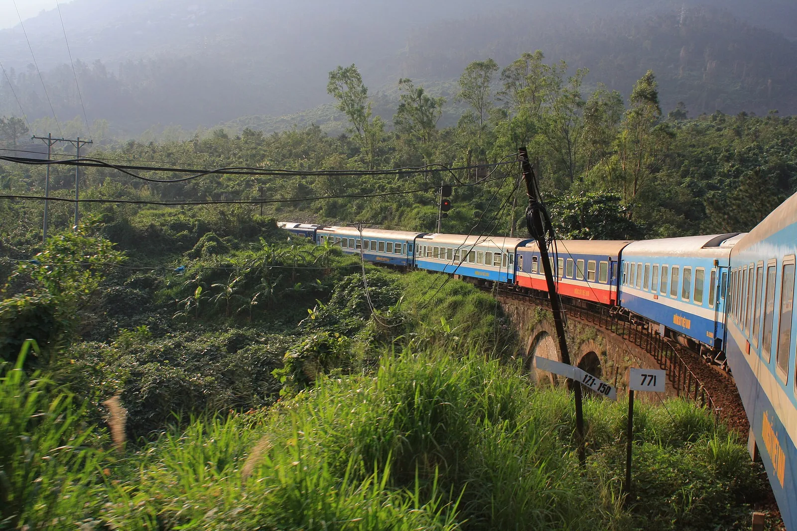 A long passenger train curving along a mountainside track through dense green vegetation on the central coast