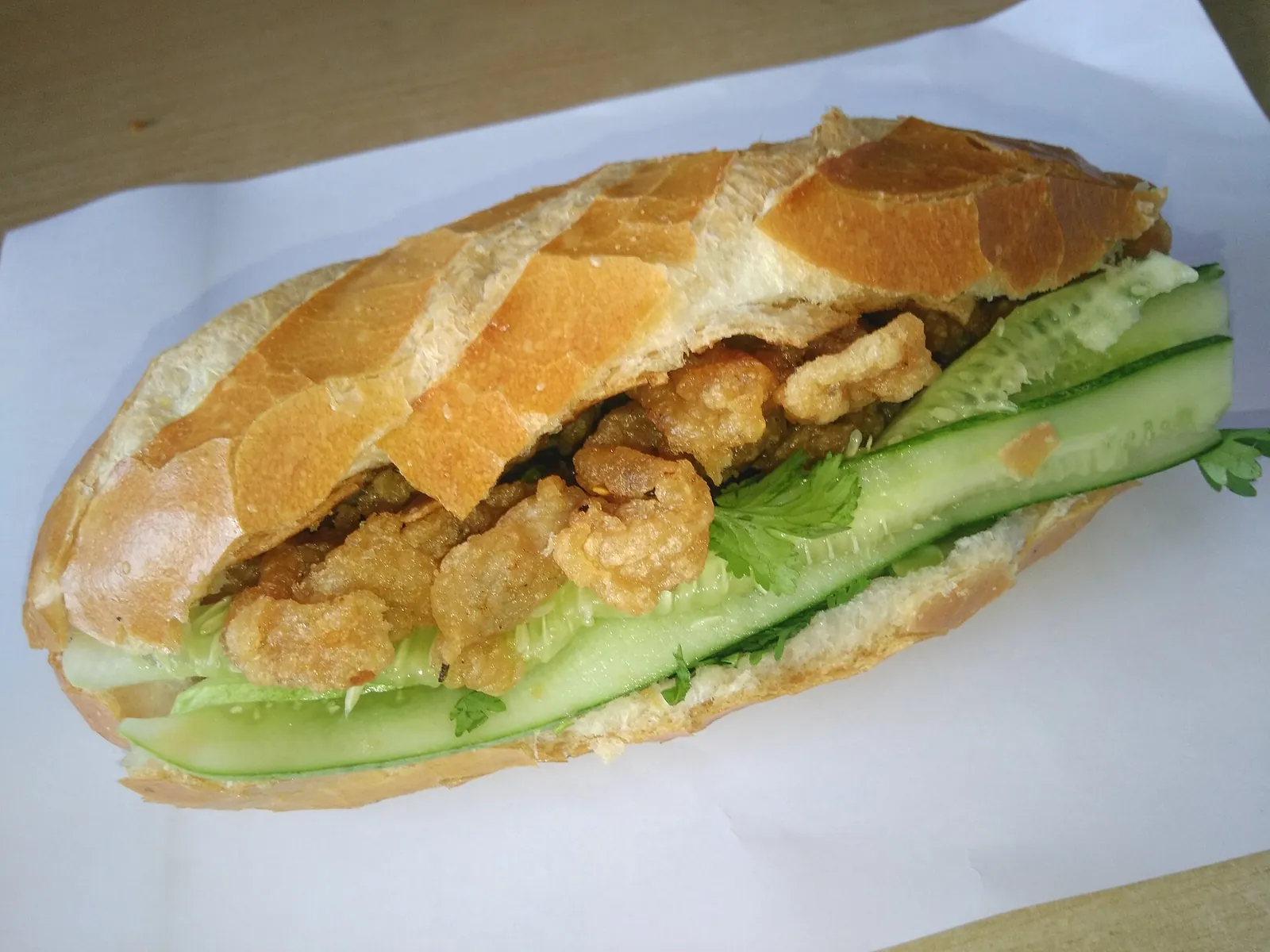 A bánh mì baguette filled with fried fish cake, cucumber and herbs on white paper