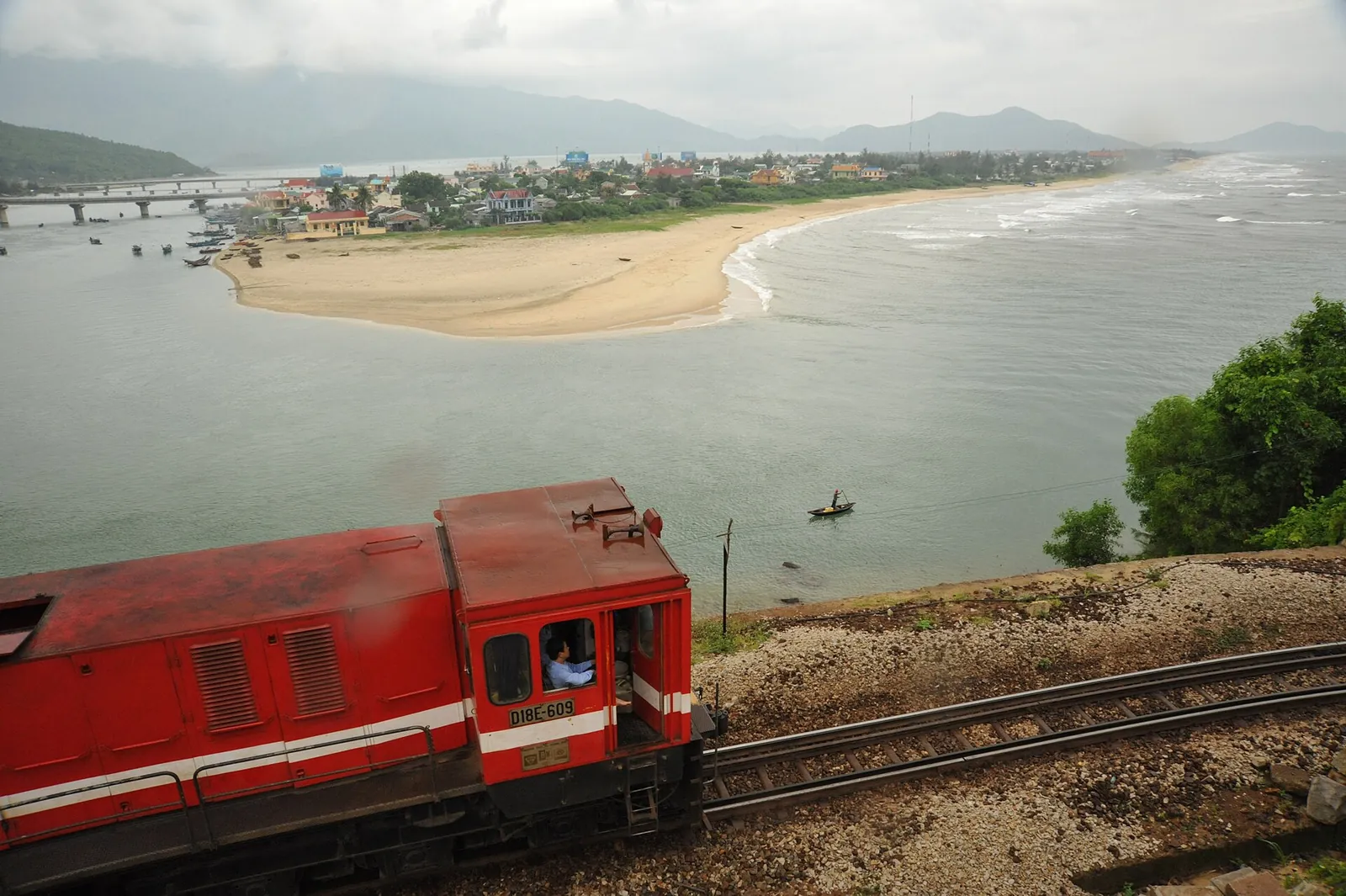 A red diesel locomotive on the railway above the lagoon mouth and beach at Lang Co