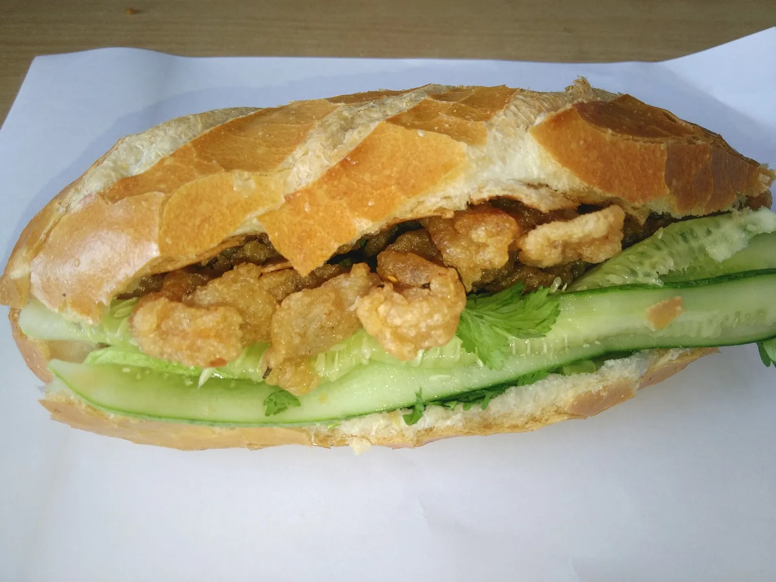 A banh mi filled with fried fish cake, cucumber and herbs on white paper