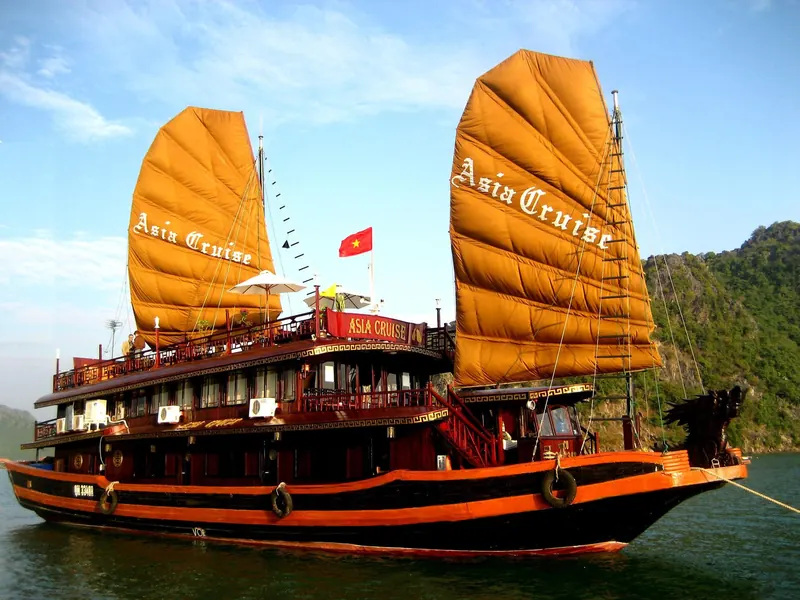 A wooden junk with orange sails moored among the limestone islands of Ha Long Bay under a blue sky