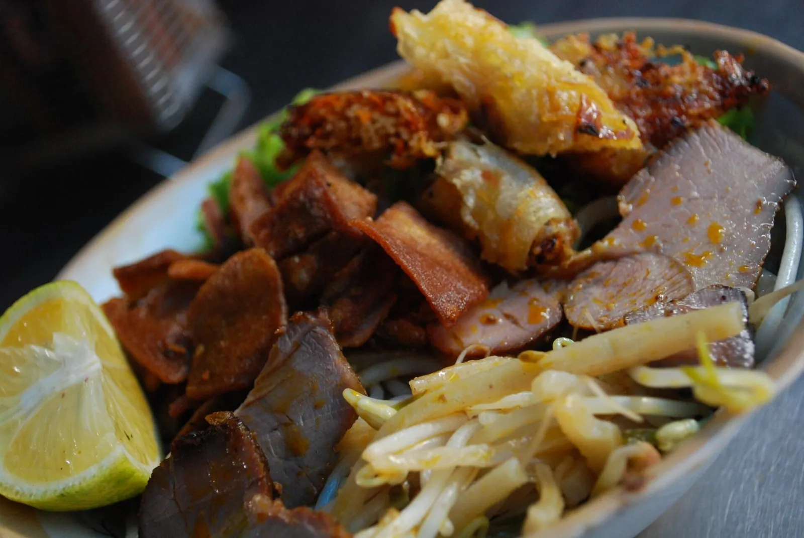 Close-up of cao lau noodles with sliced pork, crispy crackling, a fried spring roll, bean sprouts and lime