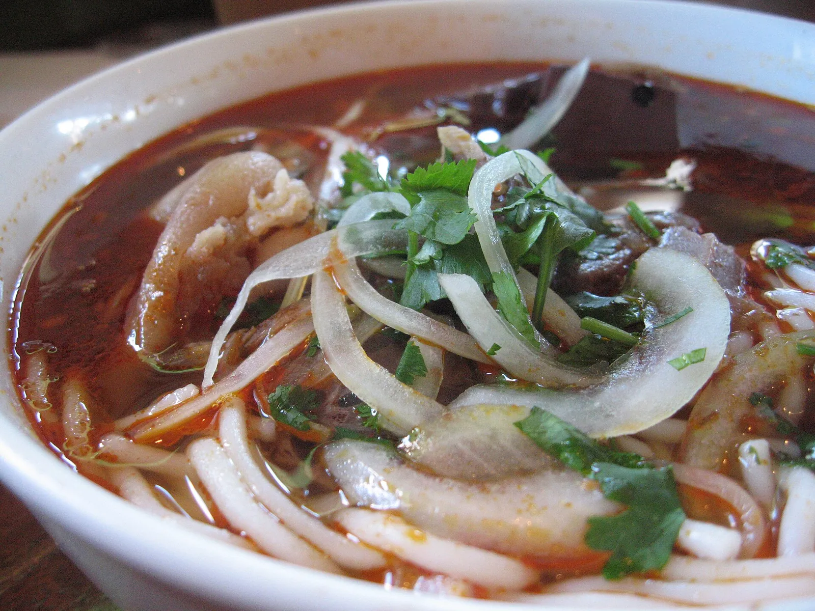 A bowl of bún bò Huế with rice noodles, sliced beef, onion and herbs in a red chilli broth