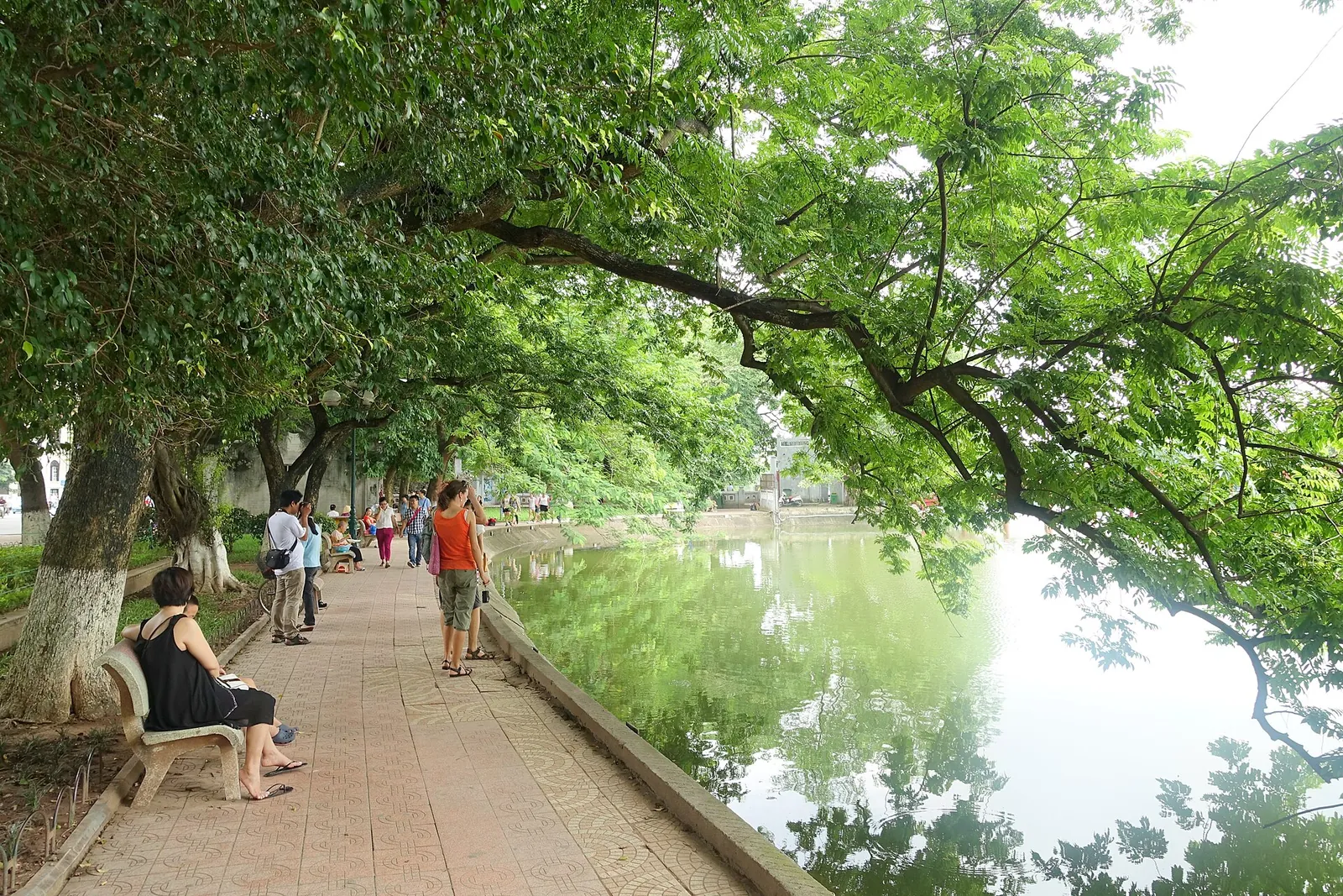 Tree-shaded lakeside path along Hoan Kiem Lake in Hanoi, with people walking and sitting on a bench
