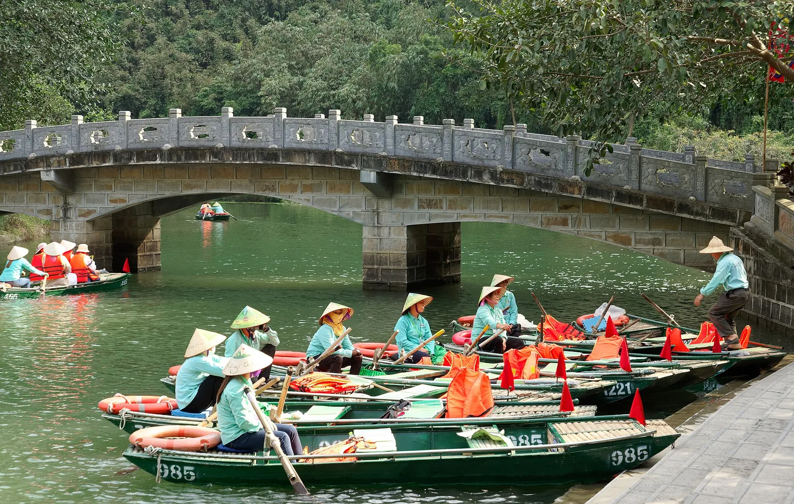 Rowers in conical hats with green rowing boats at the Trang An pier, below a stone bridge