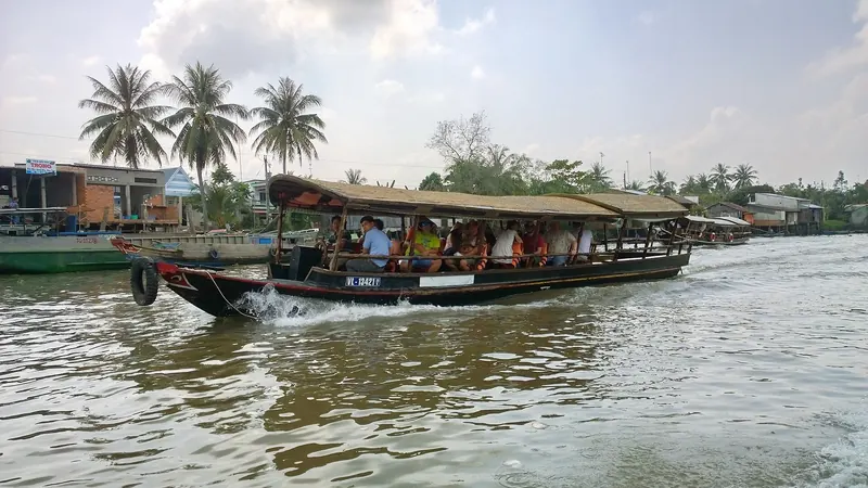 A wooden tourist boat with a covered deck crossing the brown Tien River in front of riverside houses and palms at Dong Hoa Hiep, Cai Be