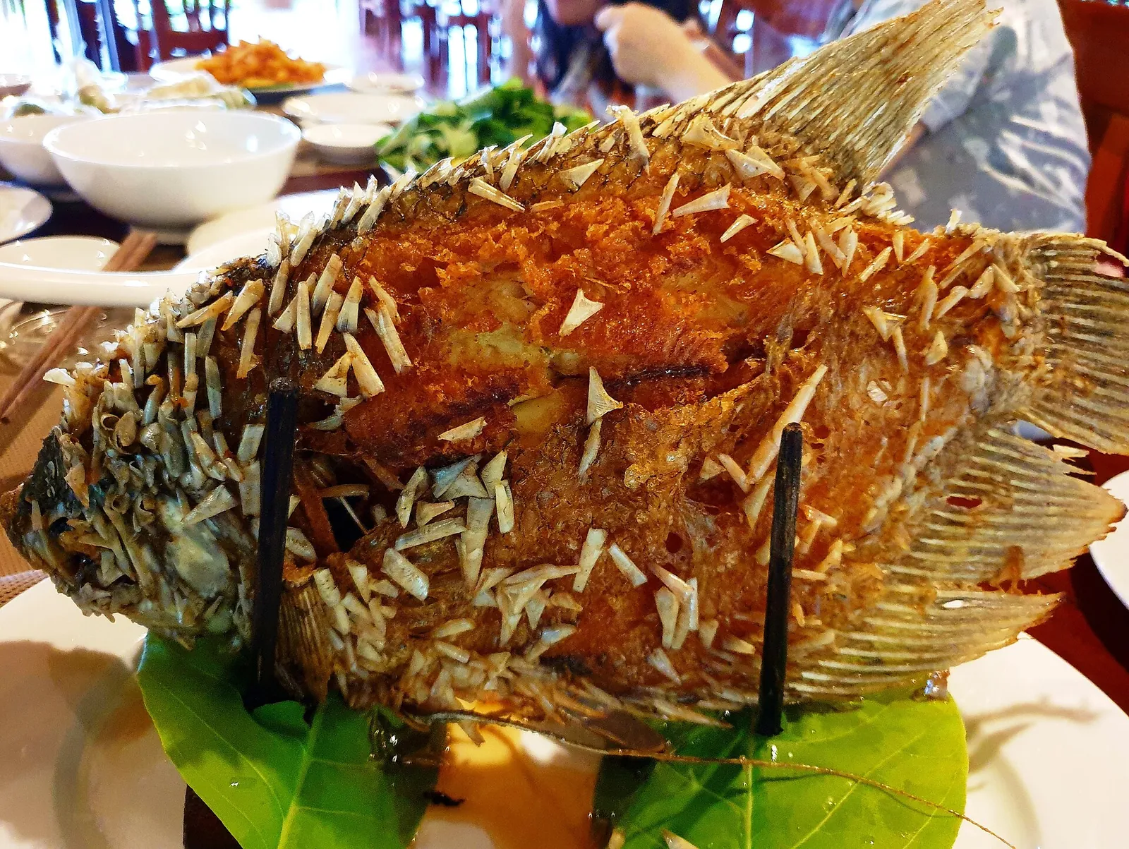 A whole deep-fried elephant-ear fish standing upright on a platter, surrounded by dishes on a restaurant table